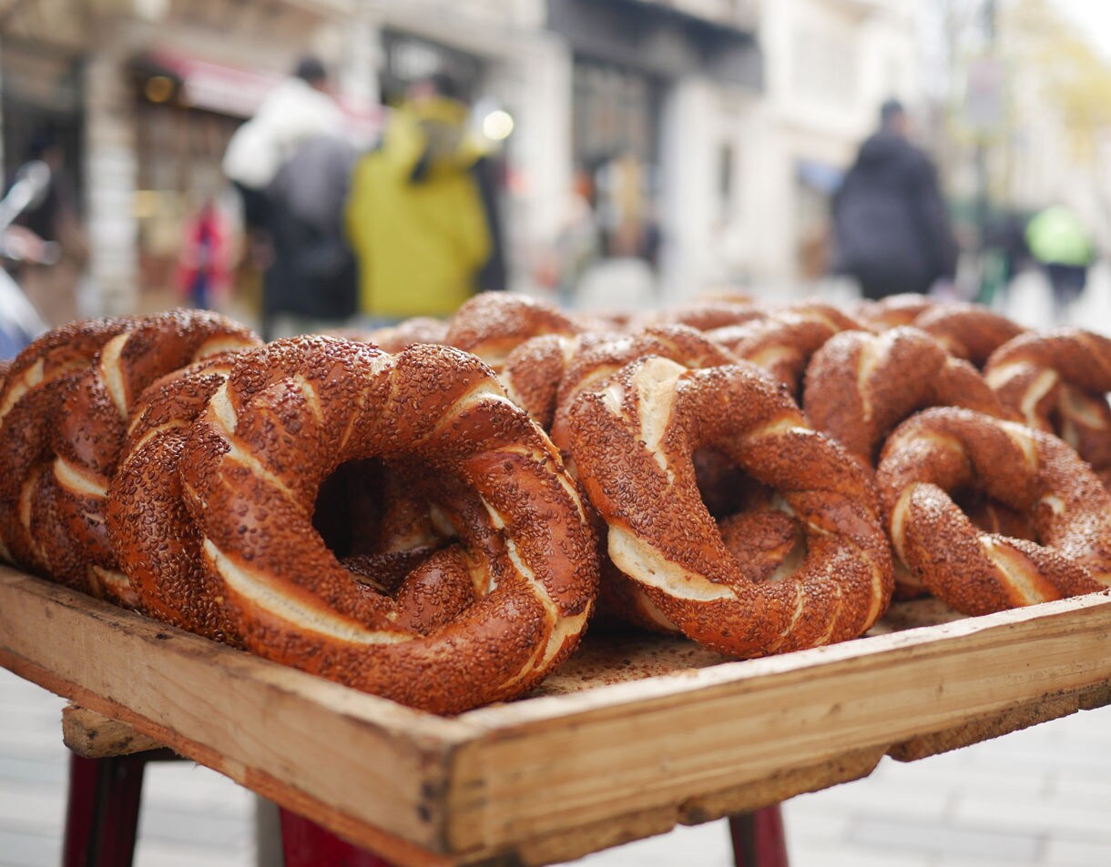 Fresh simit bread rings covered in sesame seeds displayed on a wooden tray along a busy street in Istanbul.