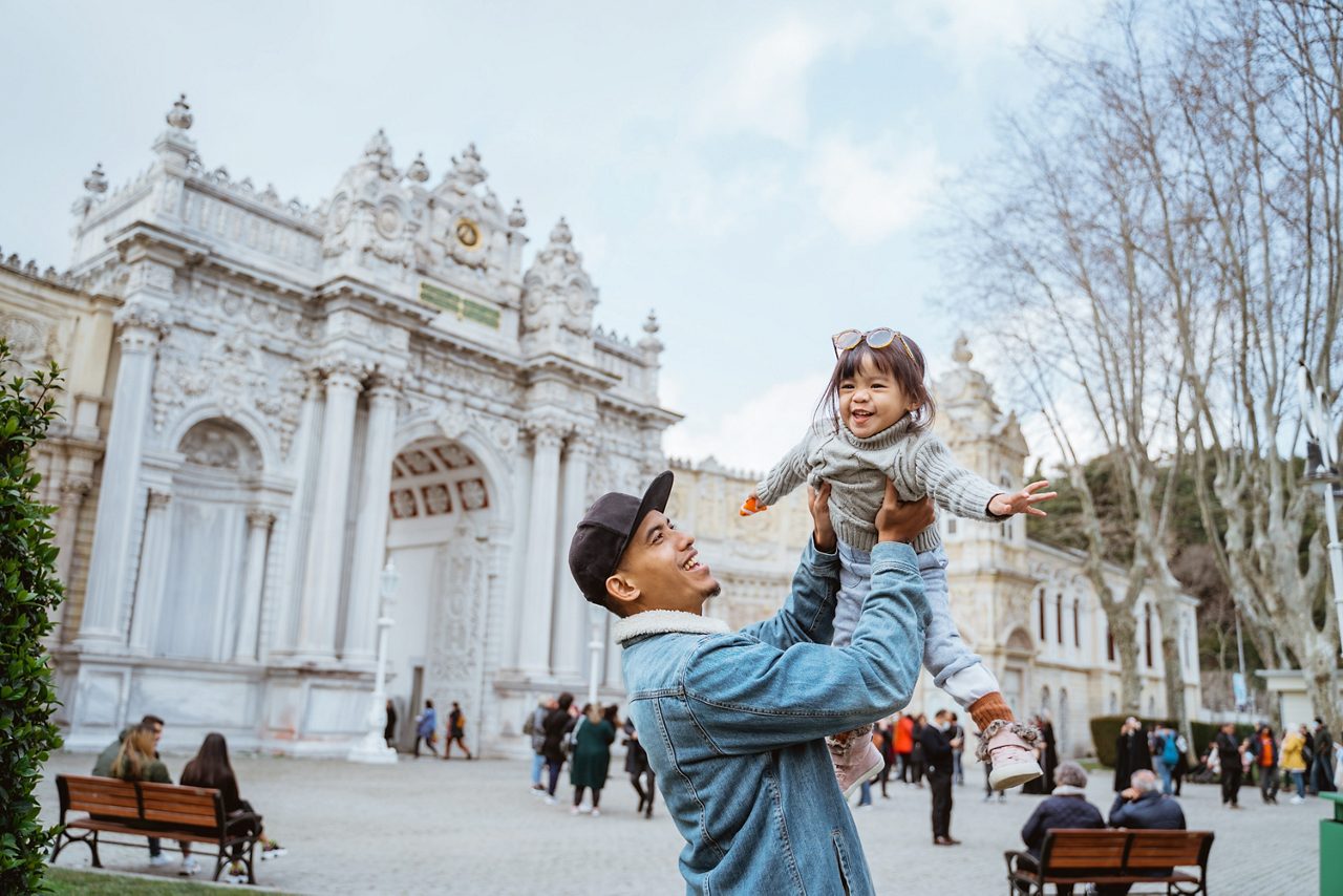 A smiling man in a denim jacket holds a laughing young girl high in the air in front of the ornate white facade and grand columns of Dolmabahçe Palace.