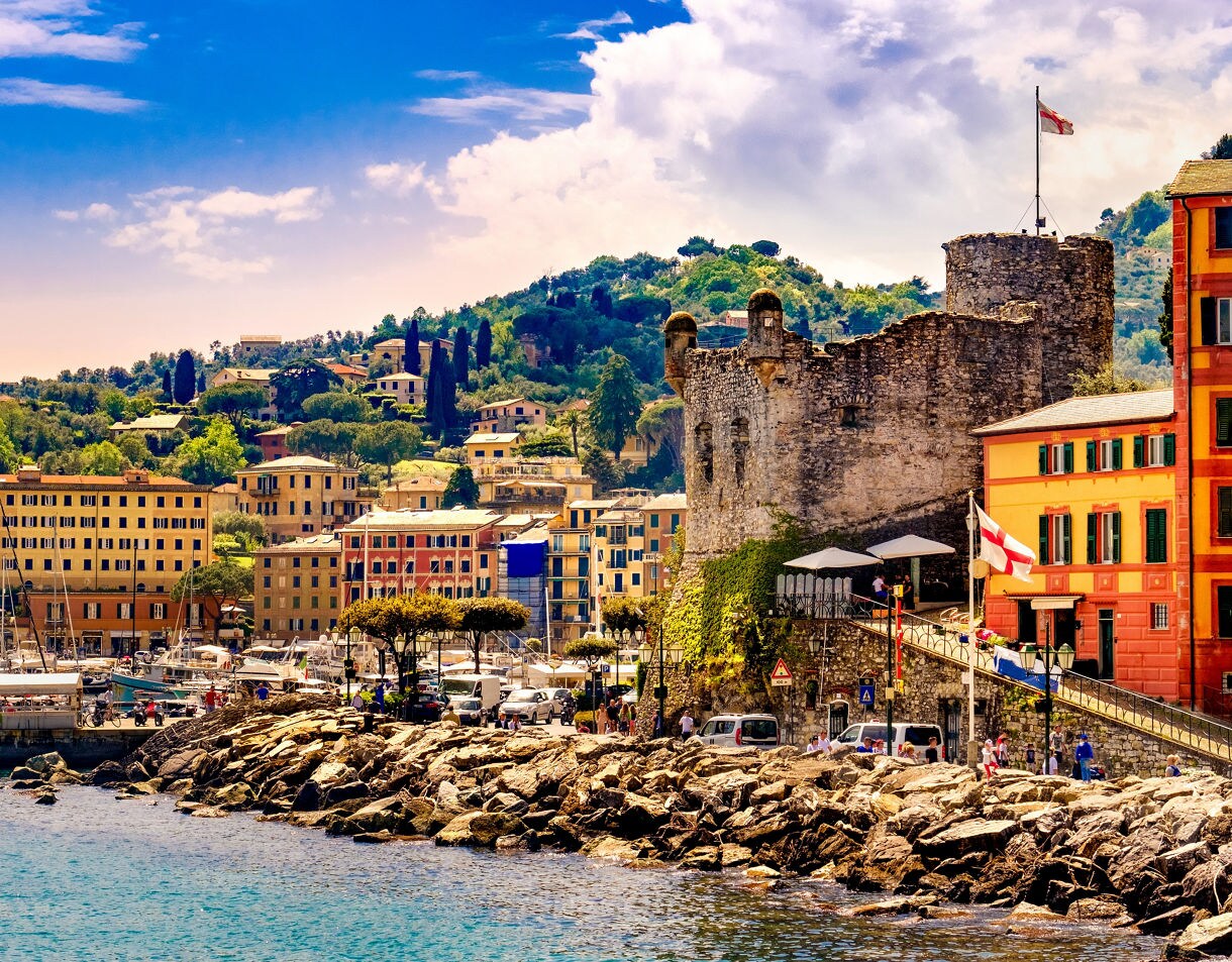 Colorful coastal town with a stone castle, boats in the harbor and hillside villas under a bright sky.