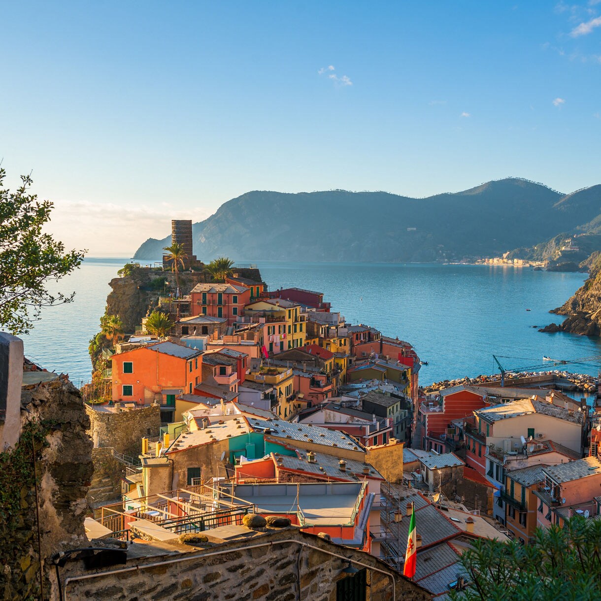 Aerial view of Vernazza in the Cinque Terre with colorful cliffside buildings, a stone watchtower and terraced hills overlooking a calm blue bay at sunset.