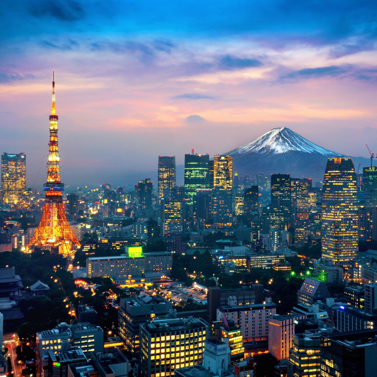 Aerial night view of the illuminated Tokyo skyline at dusk with Tokyo Tower glowing orange on the left and the snow-capped peak of Mount Fuji rising above the city under a dramatic pink and blue sky.