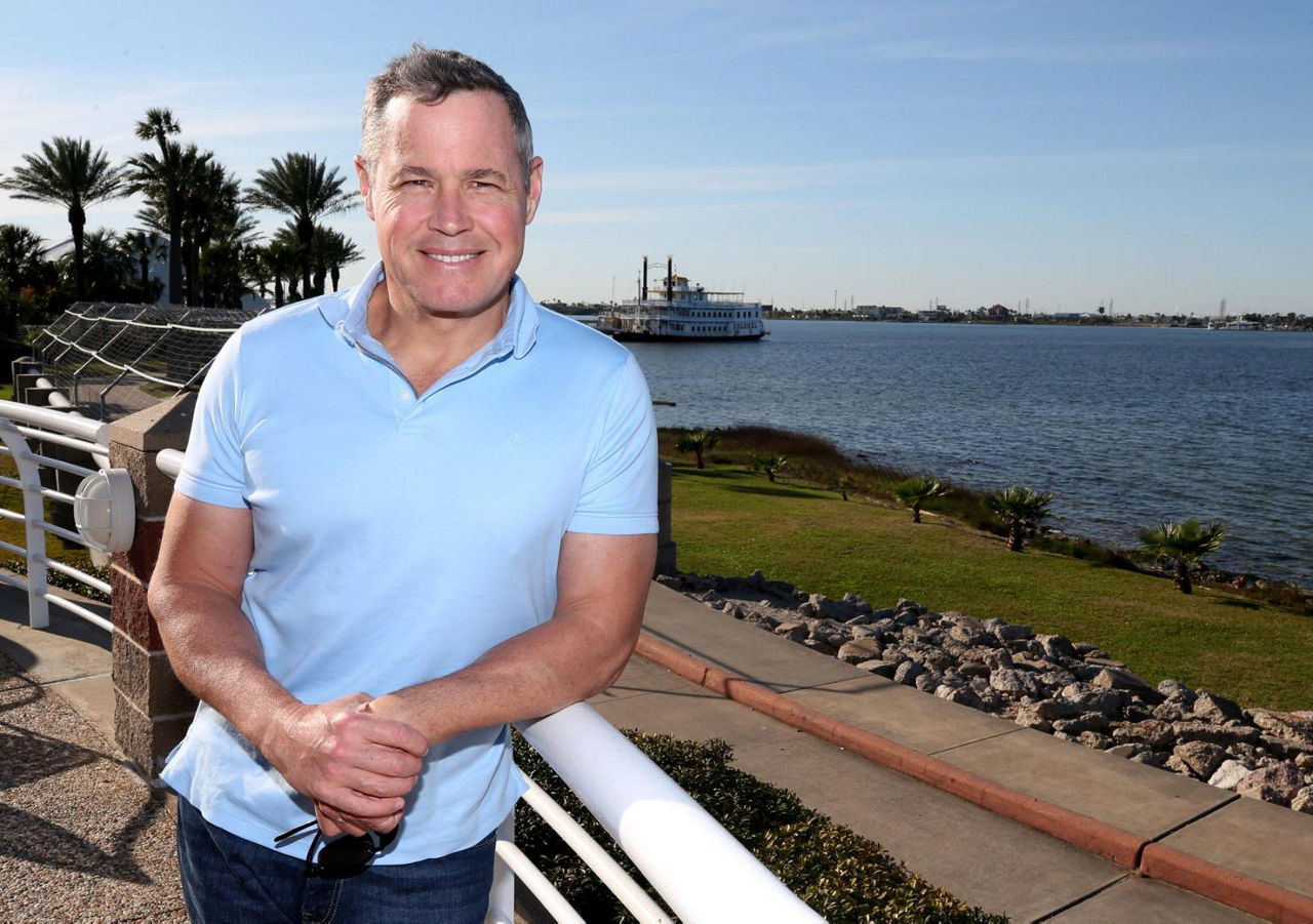  Jeff Corwin leans against a railing beside a river with a paddlewheel boat behind him.