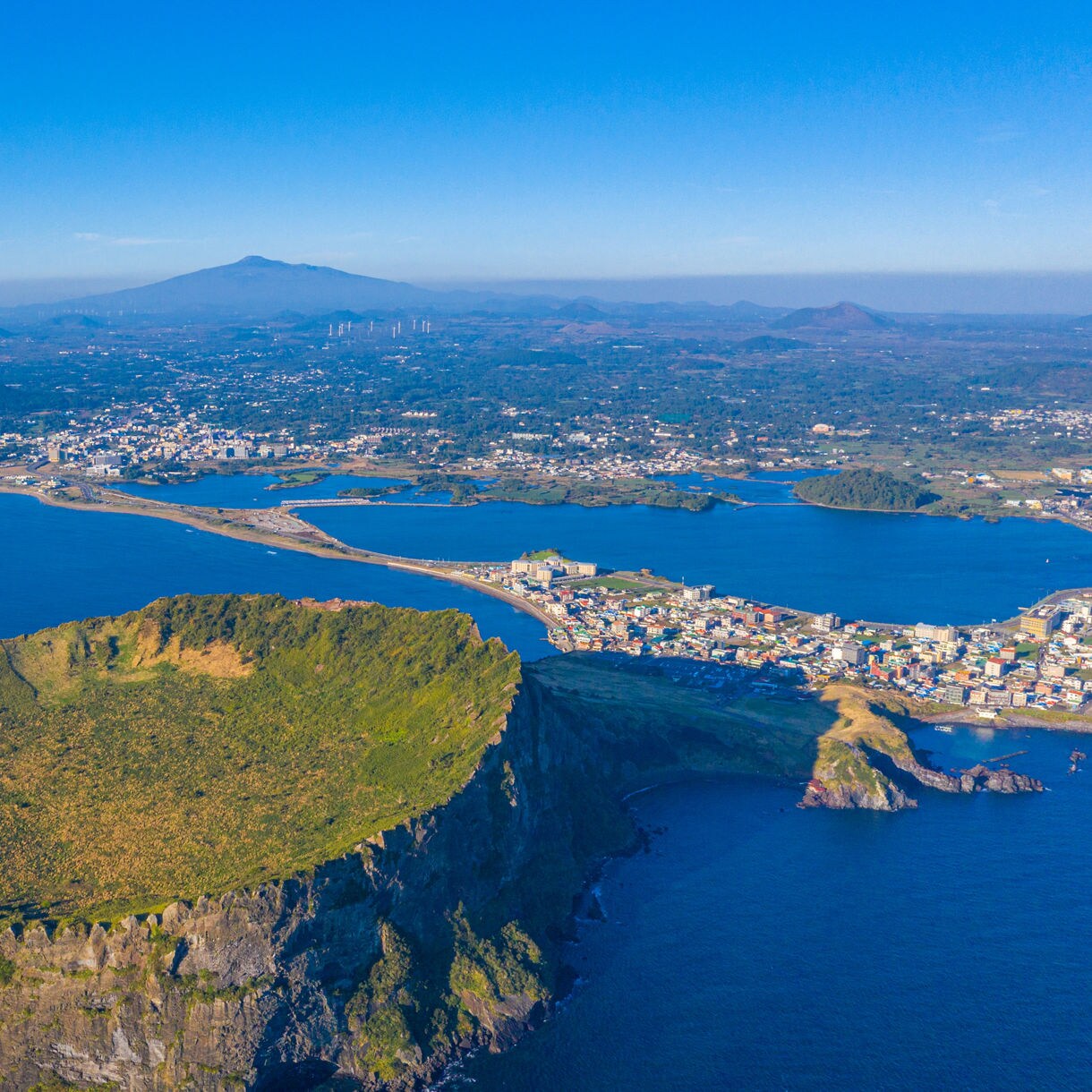 Aerial view of Seongsan Ilchulbong crater on Jeju Island, South Korea, with lush green slopes, surrounding coastline and distant mountains under clear blue skies.