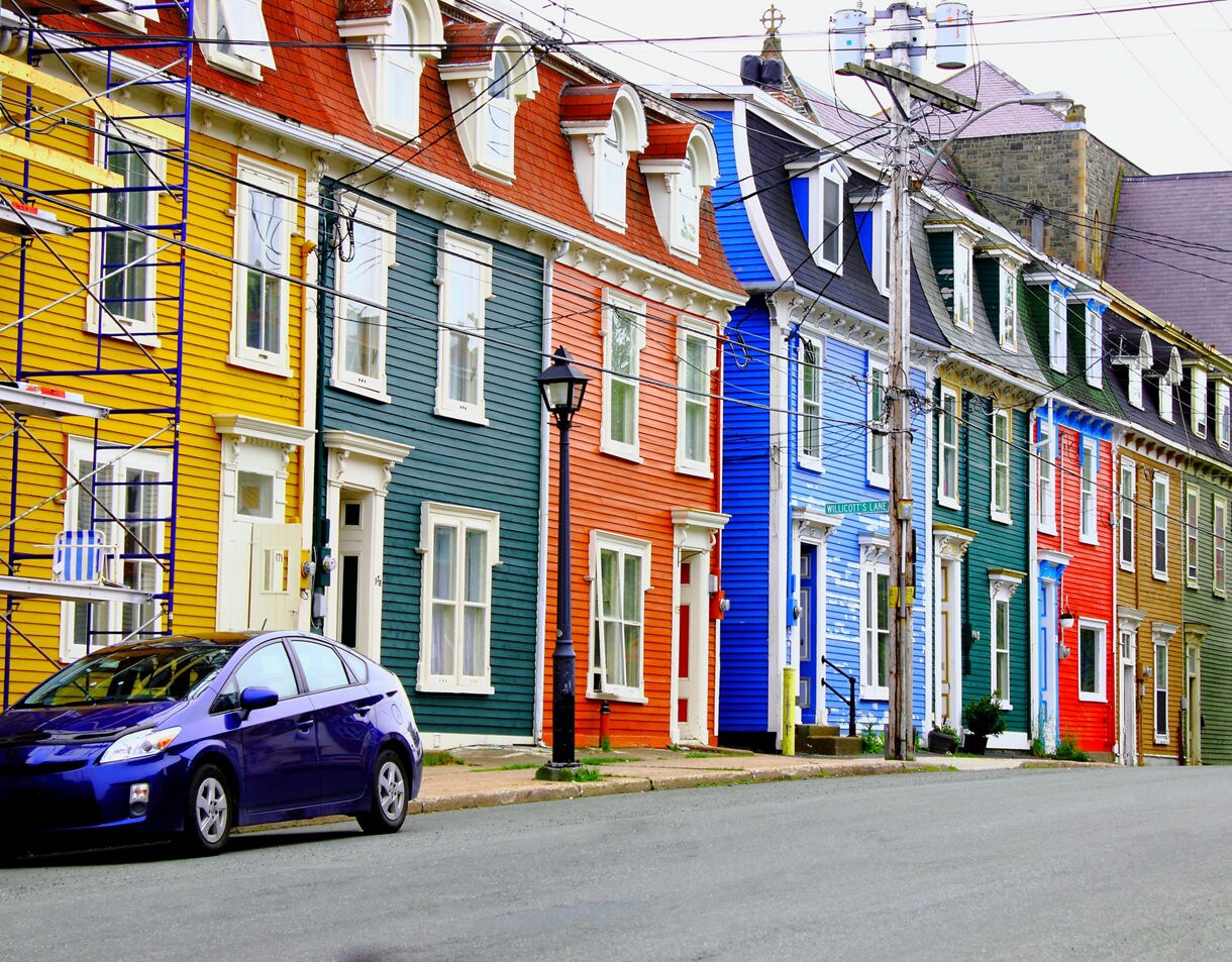 A row of brightly painted wooden townhouses in St. John’s, Newfoundland, featuring yellow, teal, orange, blue and green facades along a quiet street with cars and overhead power lines.