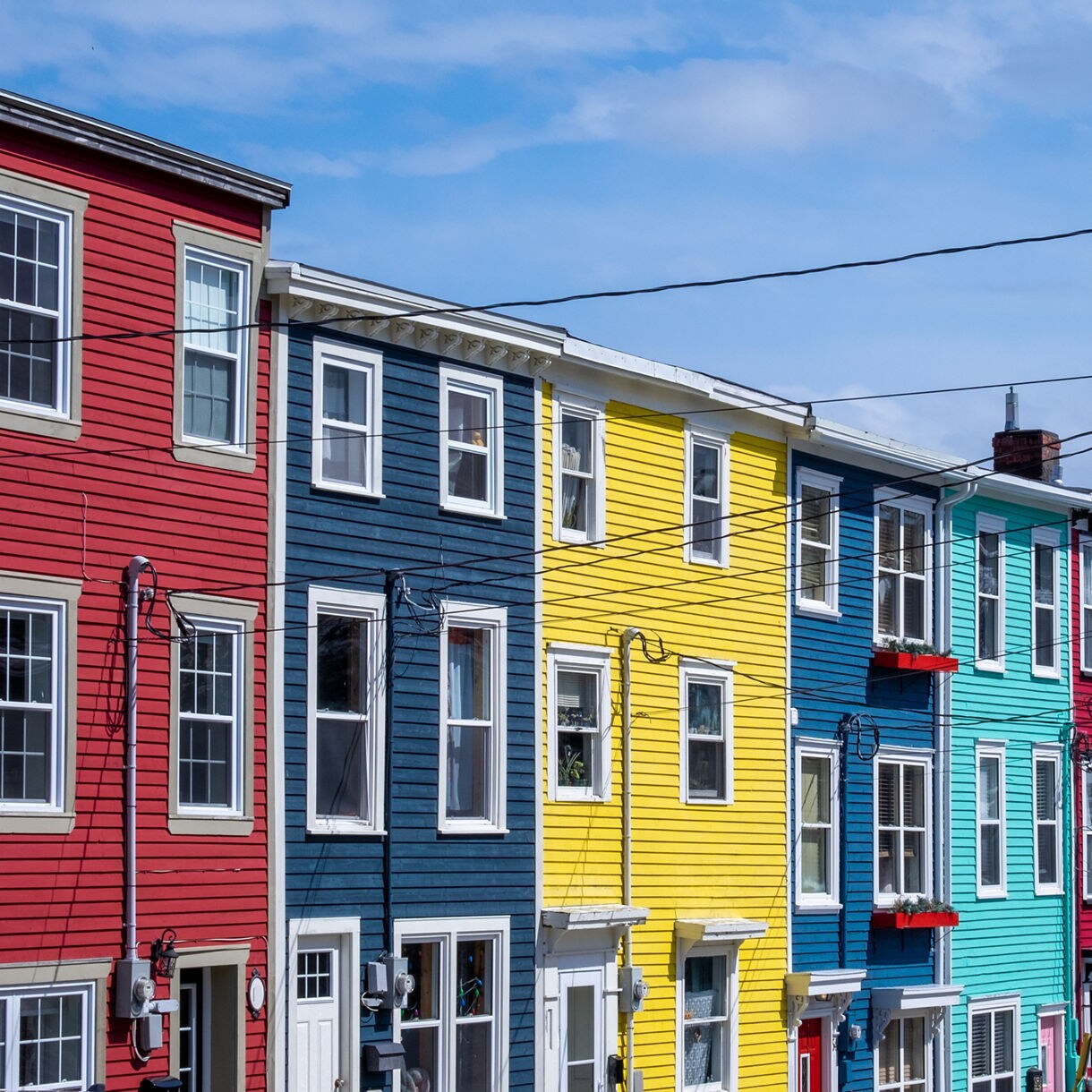A row of brightly painted wooden townhouses in St. John’s, Newfoundland, featuring red, navy, yellow, teal and green siding under a blue sky with overhead power lines.