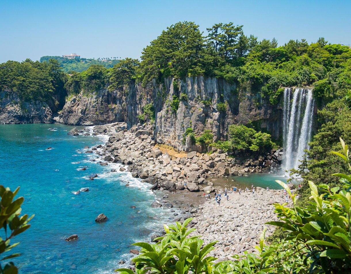  Jeongbang Waterfall on Jeju Island, South Korea, with water streaming over cliffs covered in lush greenery and flowing into the turquoise ocean below.