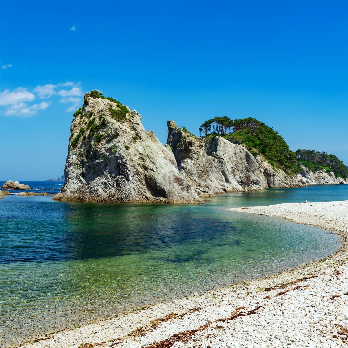 Rocky coastal inlet with tall white limestone formations rising from clear blue-green water, bordered by a curved shoreline of white pebbles under a bright blue sky.