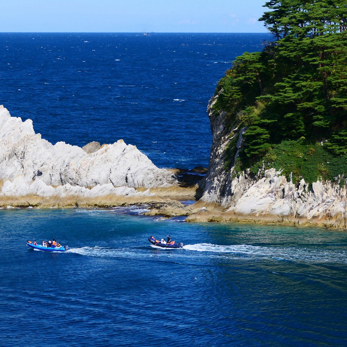 Two small sightseeing boats travel through bright blue water near Jodogahama, passing between sharp white rock formations and a forested cliff.