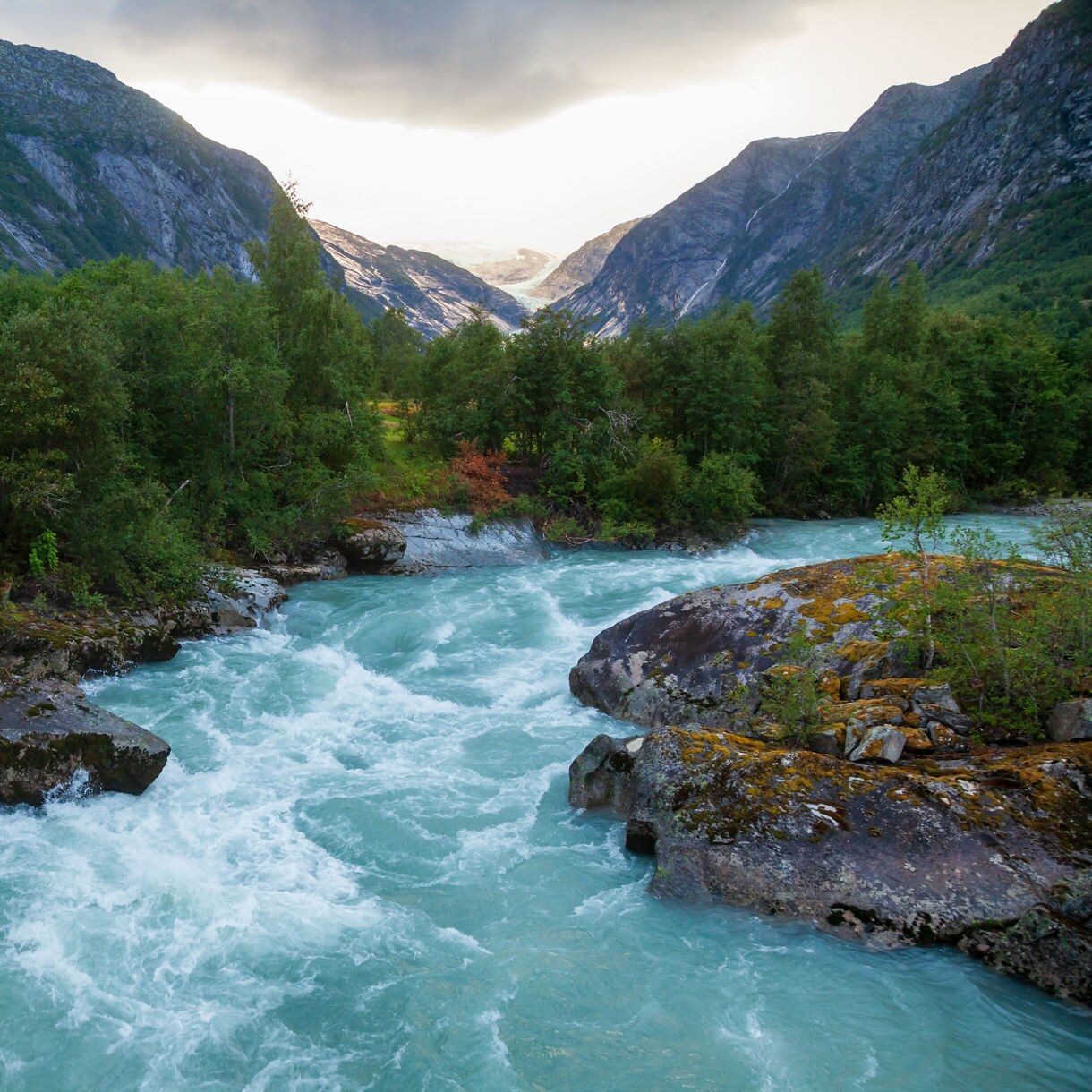 Turquoise glacial river rushing through rocky terrain and forested slopes in Jostedal Glacier National Park, Norway, with distant ice fields visible between mountain peaks.