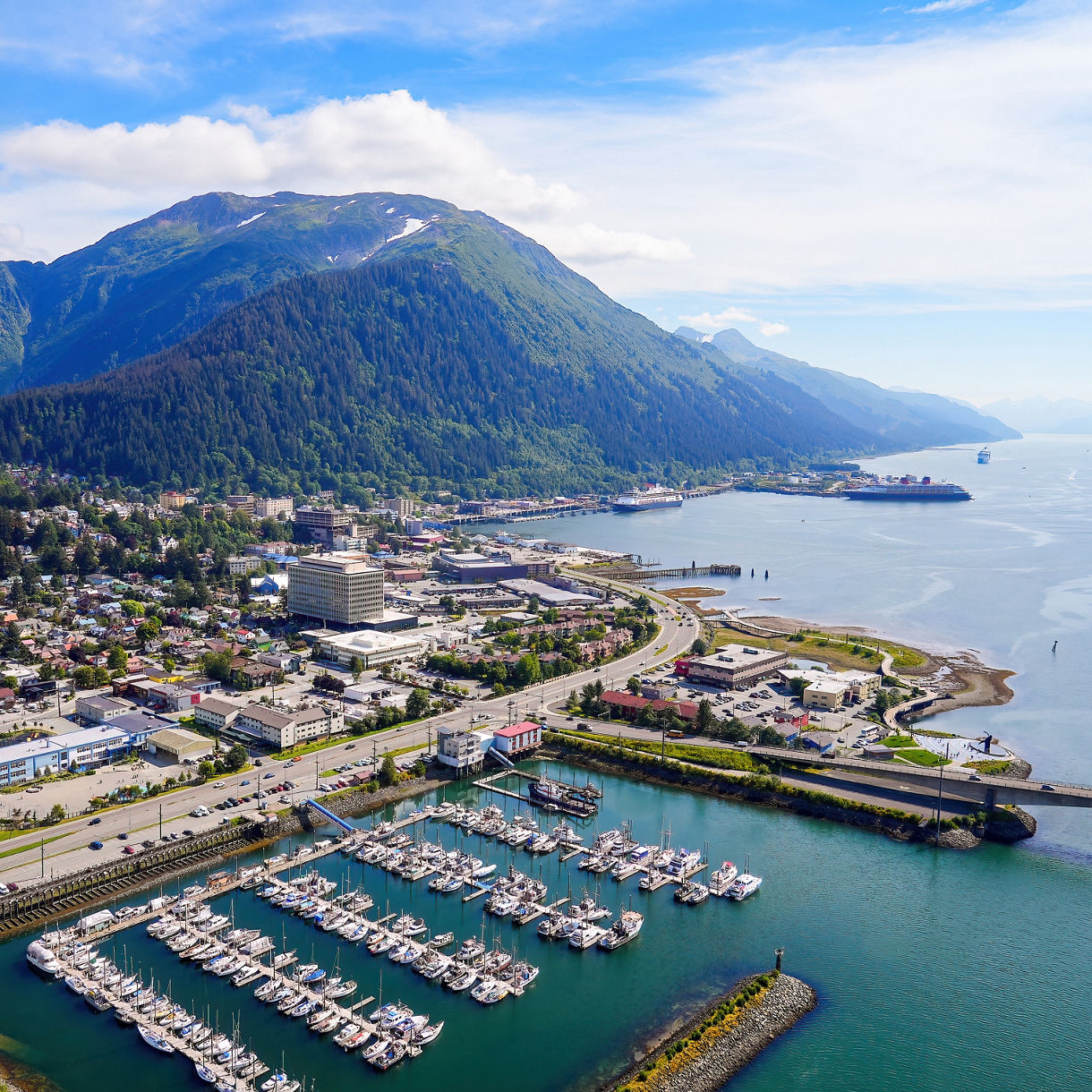 An aerial view reveals a coastal city nestled at the base of massive green mountains. A large marina packed with white boats sits in the foreground next to clear blue waters.