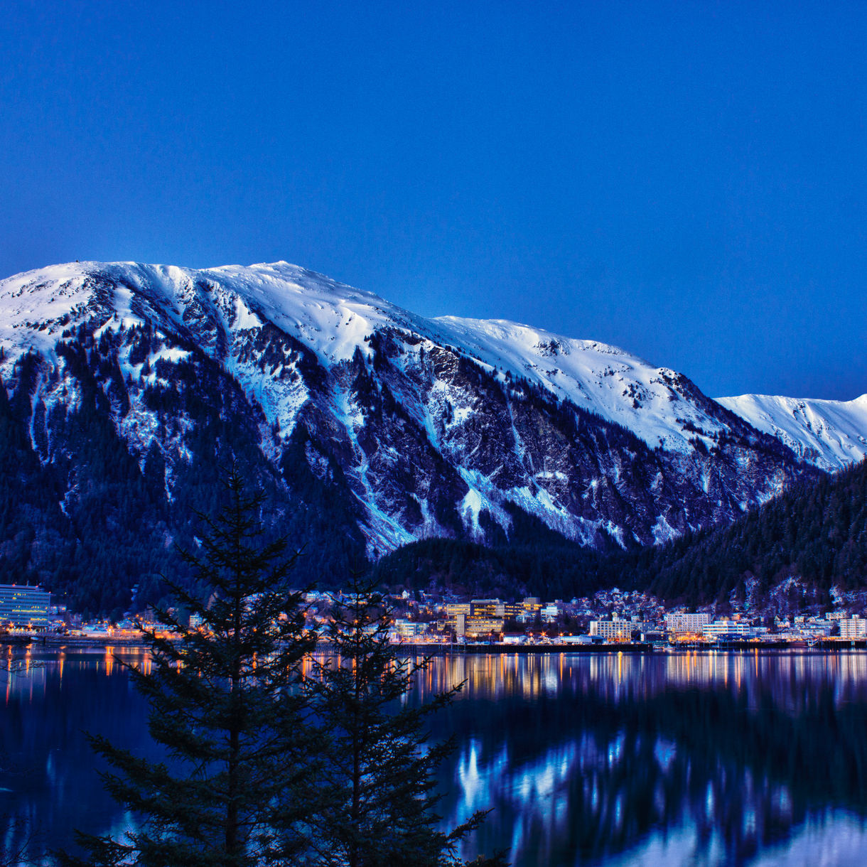 A snow-covered mountain rises behind a small town nestled along the edge of a calm lake. The scene is captured at twilight, with deep blue sky and subtle city lights glowing at the shoreline. Forested slopes frame the town, and the water reflects the mountains and lights like a mirror. The overall atmosphere is serene, cold, and peaceful in a winter setting.
