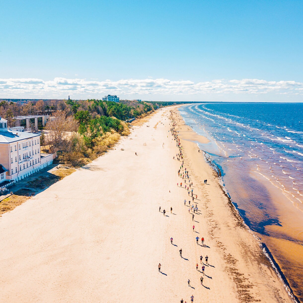 Aerial view of Jūrmala beach in Latvia, with people walking along the wide sandy shoreline beside calm blue waves and forested areas near seaside villas.