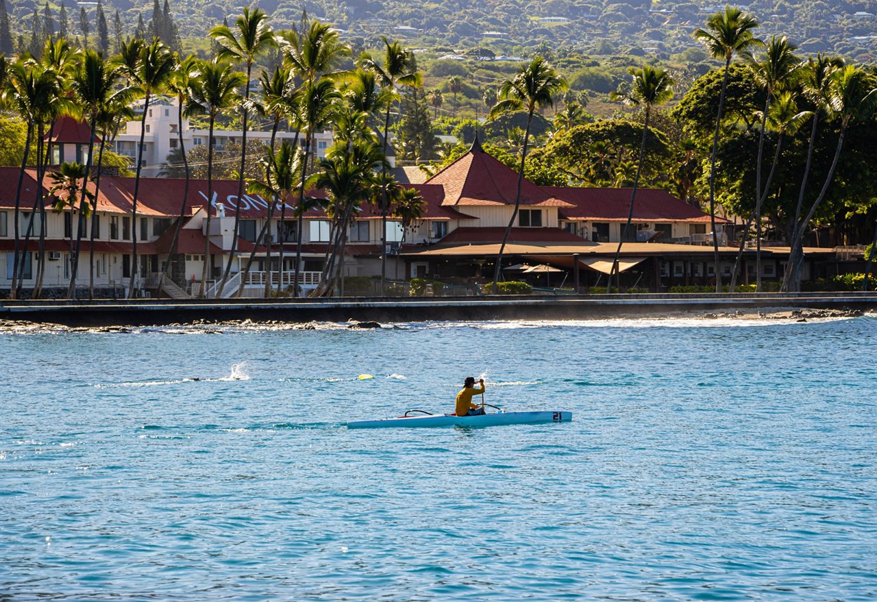 Person paddling an outrigger canoe in Kailua Bay with palm trees and red-roofed beachfront buildings in the background.