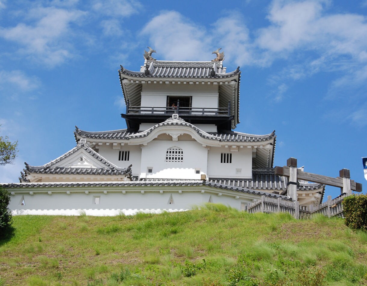 Traditional Japanese castle with white plaster walls and gray tiled roofs standing on a grassy hill, featuring ornate gables and decorative rooftop figures under a vivid blue sky.