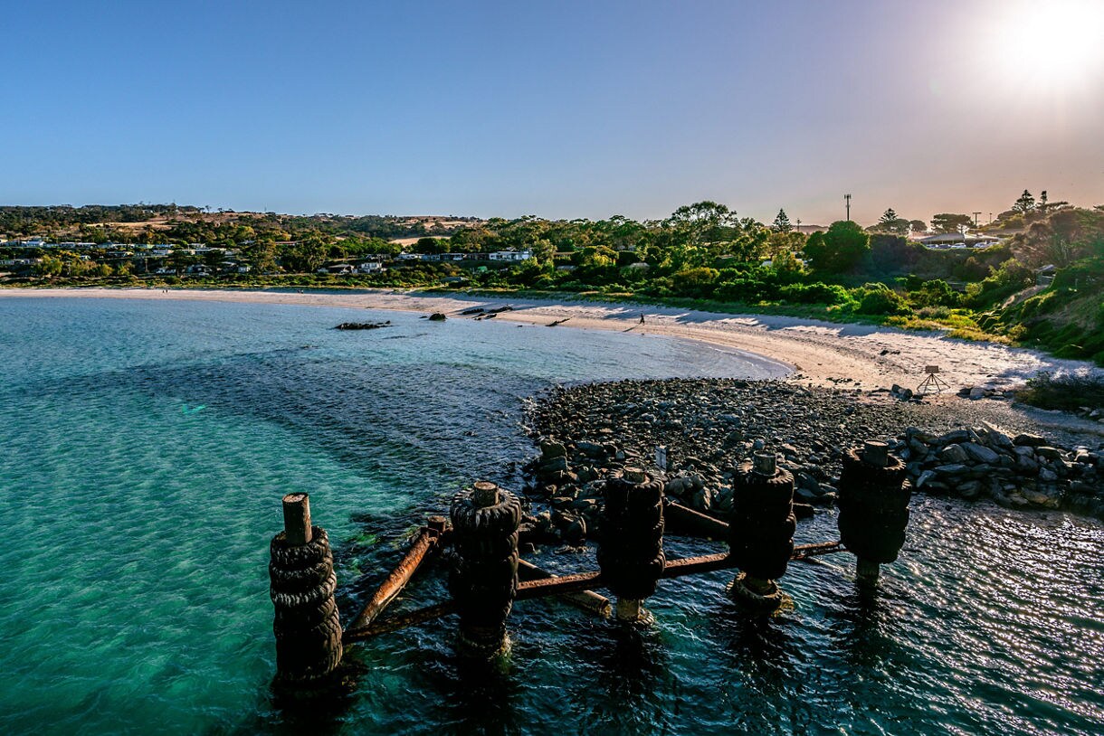 Aerial view of a sunlit beach on Kangaroo Island with turquoise water, rocky shoreline and remnants of an old pier.