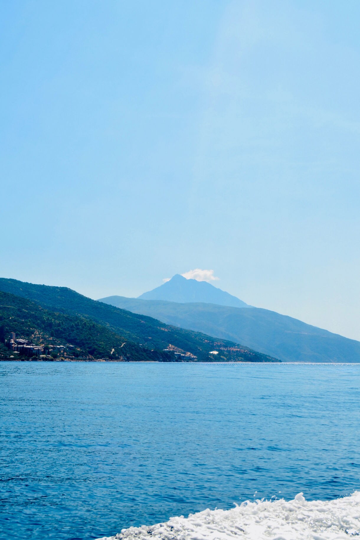 A serene coastal scene showing deep blue water, rolling green hills, and a tall mountain peak in the hazy distance, with gentle boat wake in the foreground.