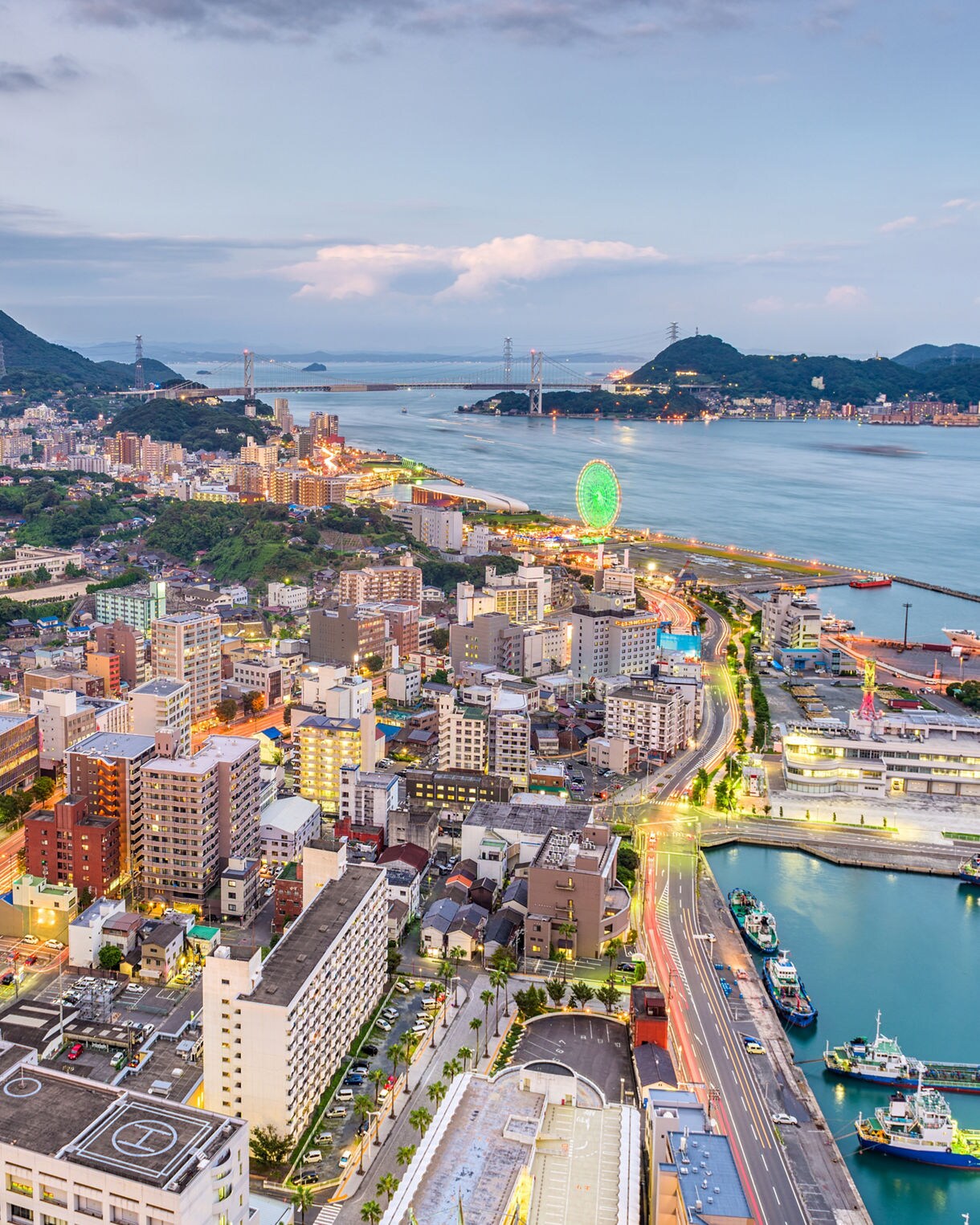 Elevated view of Shimonoseki at dusk showing illuminated city streets, a glowing green Ferris wheel, and the Kanmon Bridge spanning calm blue water between forested hills.