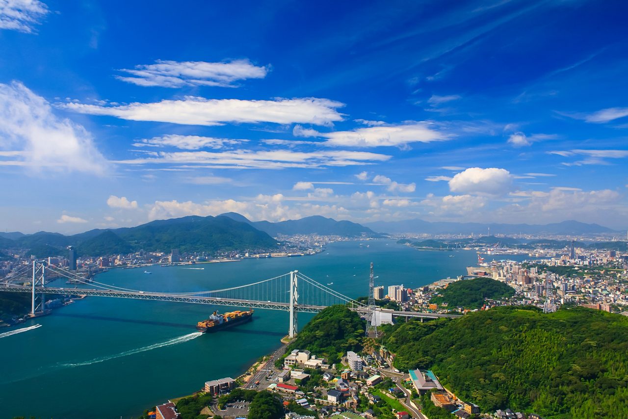 A wide aerial view of the Kanmon Strait featuring a large suspension bridge, a cargo ship, densely built coastal cities, and green mountains under a bright blue sky with streaked clouds.