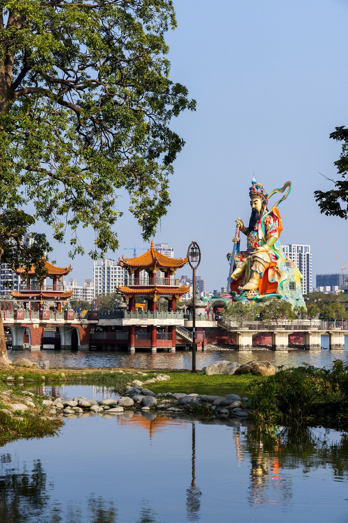 View of the colorful Guandi statue and traditional pagodas beside Lotus Pond in Kaohsiung, Taiwan, with city buildings in the background and calm water in the foreground.