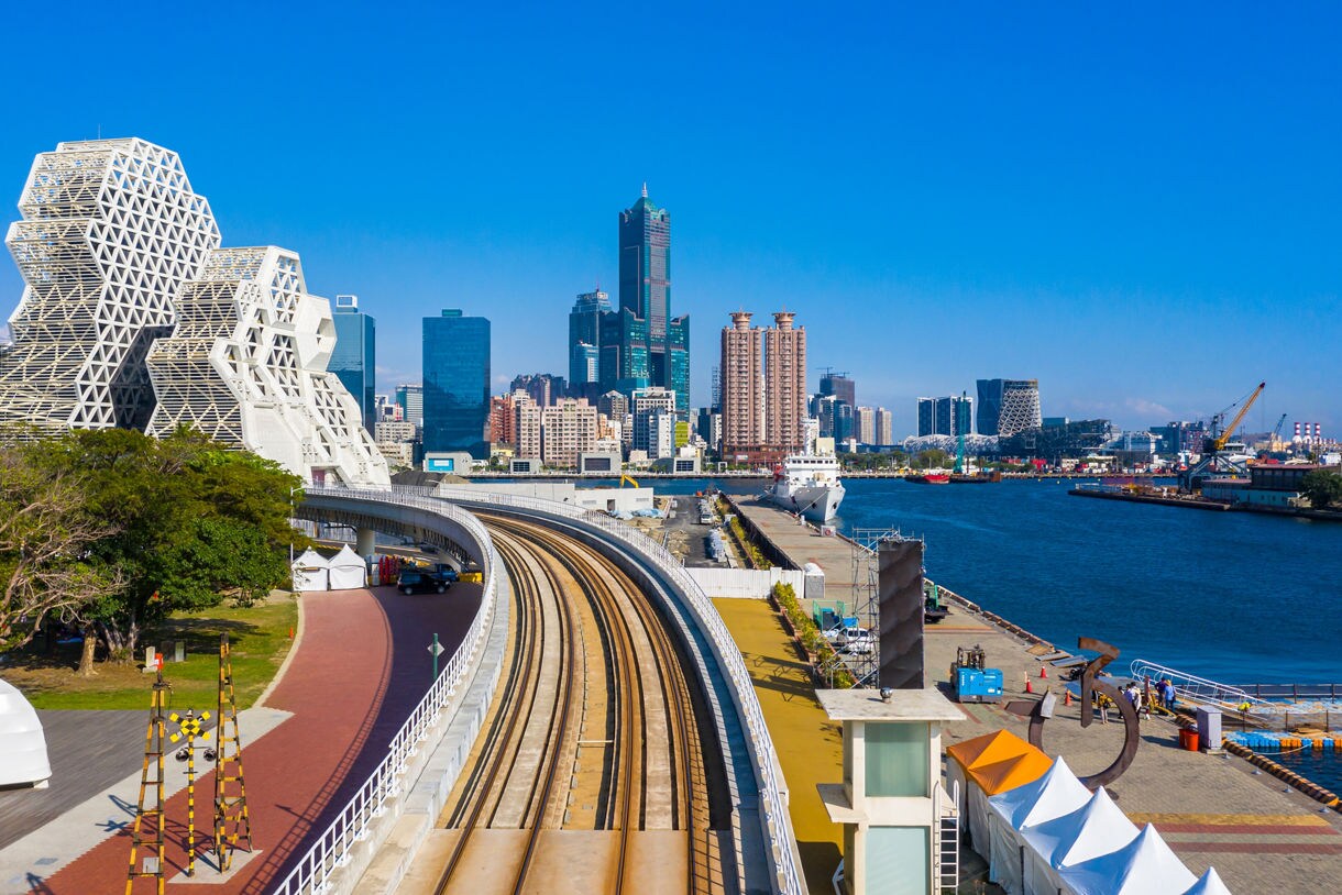 View of Kaohsiung, Taiwan, featuring the curved white Kaohsiung Maritime Cultural Center, urban skyscrapers and waterfront docks under a bright blue sky.
