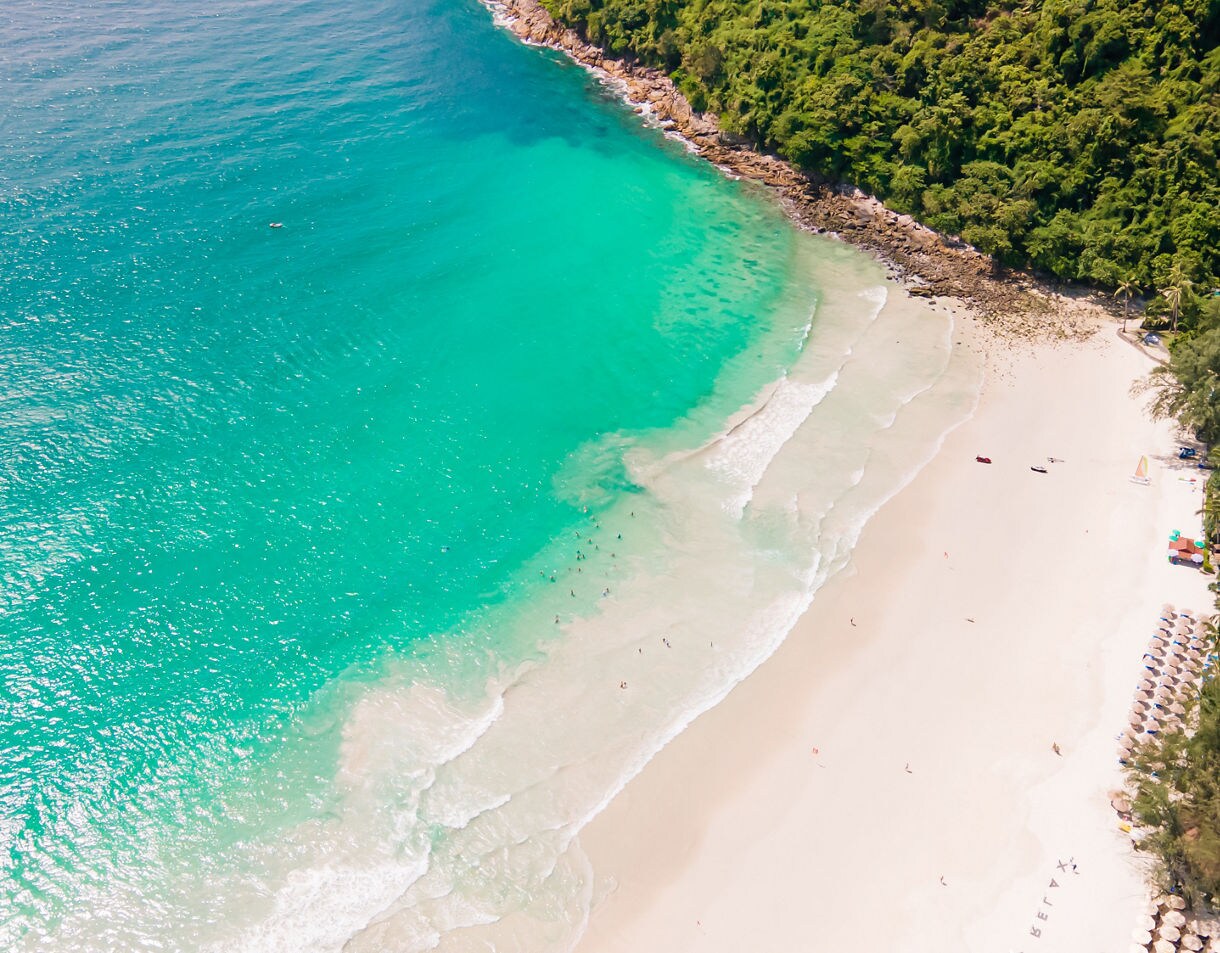 Aerial view of Karon Noi Beach in Phuket with white sand, turquoise waves and green hills bordering the shoreline.