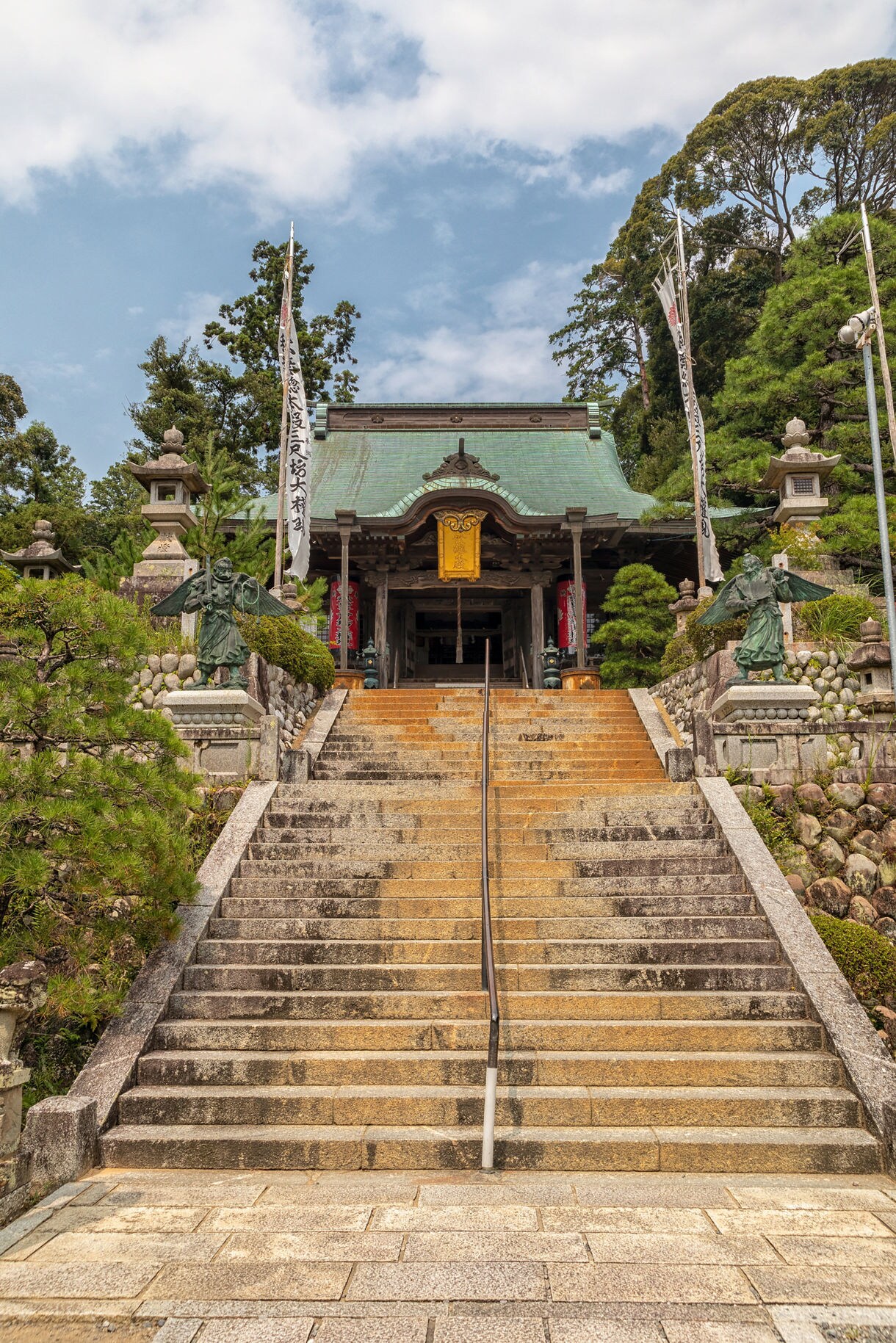 Wide stone staircase leading up to a historic Japanese temple with a green-tiled roof, flanked by statues, stone lanterns and lush trees under a partly cloudy sky.