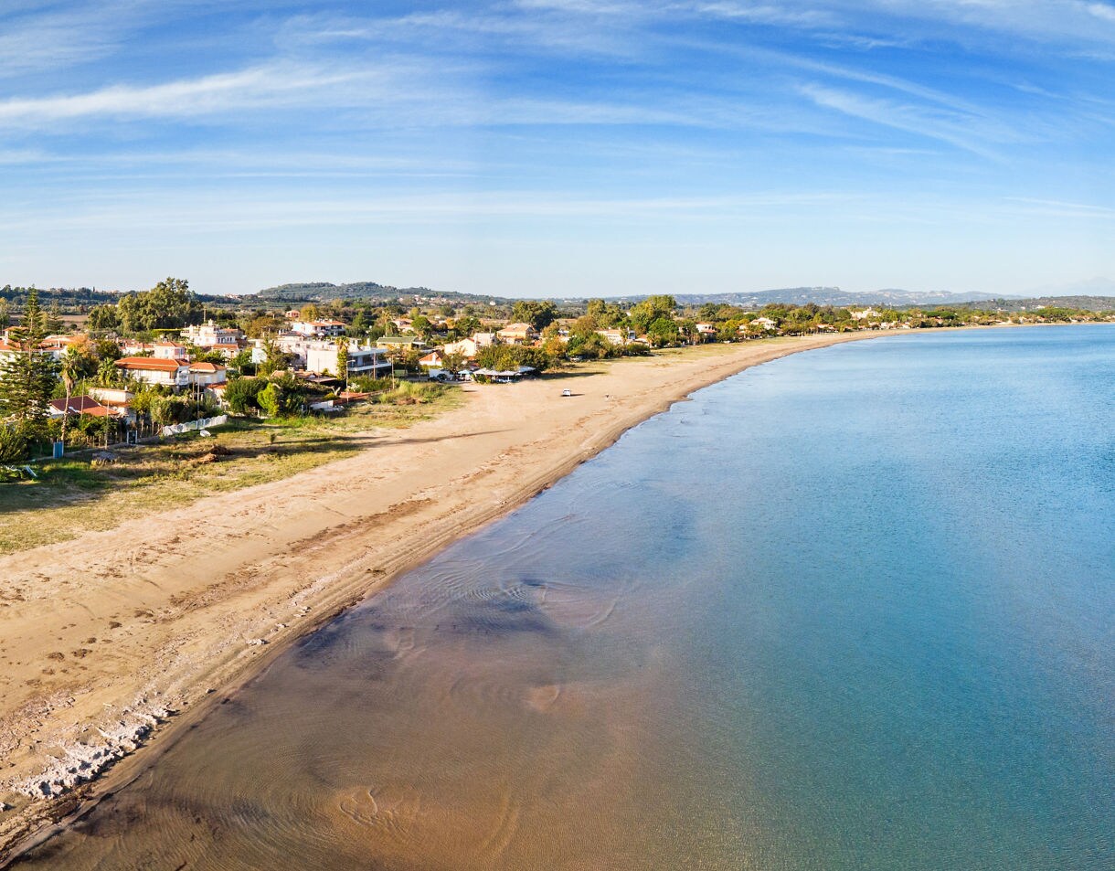 Aerial view of Katakolon Beach in Greece, with golden sand curving along the shoreline and calm blue waters stretching toward a coastal village.