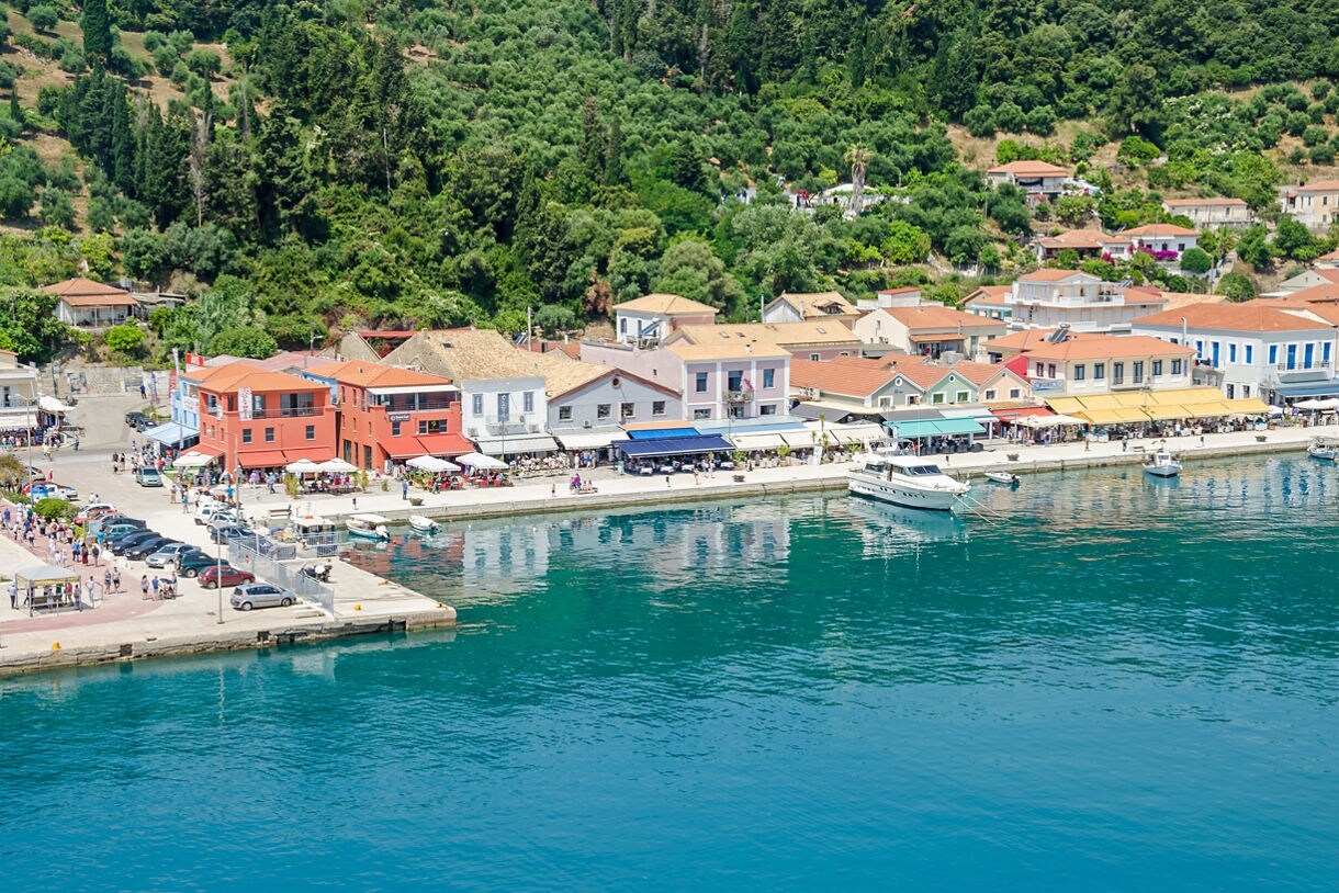 Waterfront view of Katakolon, Greece, featuring pastel-colored buildings, boats docked along the pier and lush green hills rising behind the town.
