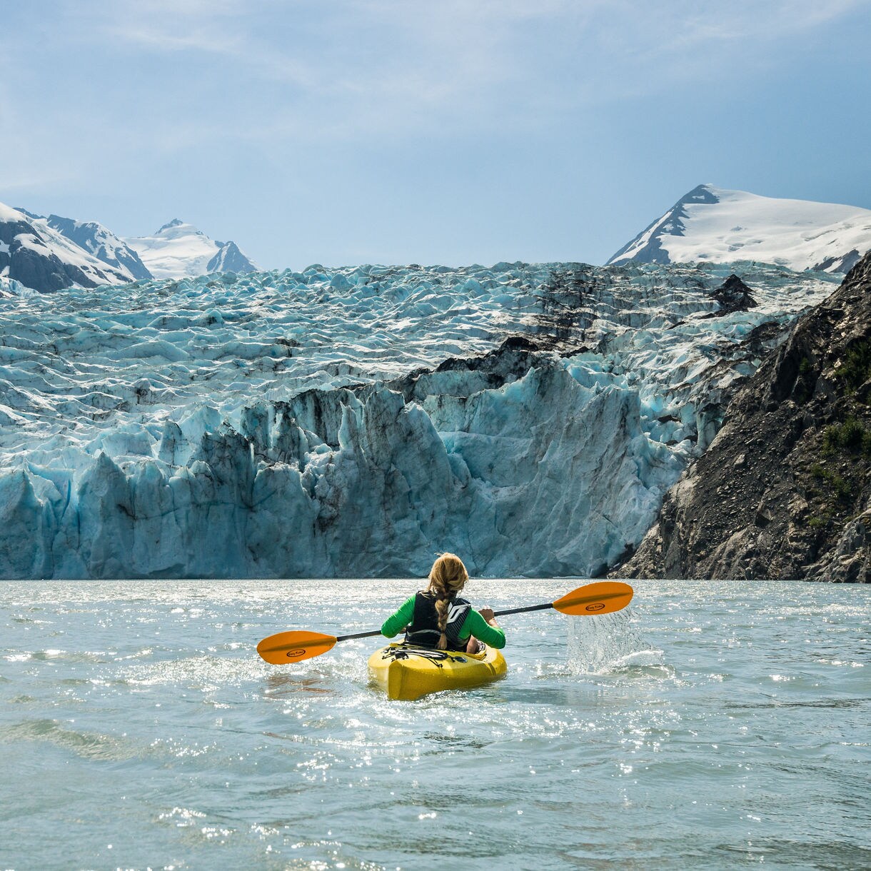 A woman in a yellow kayak paddles toward the jagged blue-white face of a massive glacier in Alaska, flanked by rocky cliffs and snow-capped mountains under a bright blue sky.