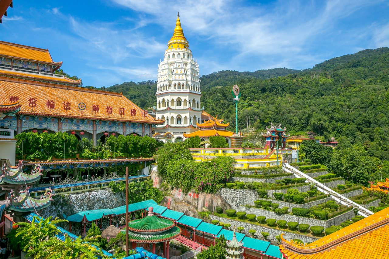 The Kek Lok Si Temple, a renowned Buddhist site, is surrounded by vibrant greenery and tiered gardens. The golden spire of the pagoda stands out against the blue sky, creating a serene and picturesque view. Traditional architecture and intricate details are visible throughout the temple complex, nestled in a mountainous landscape.