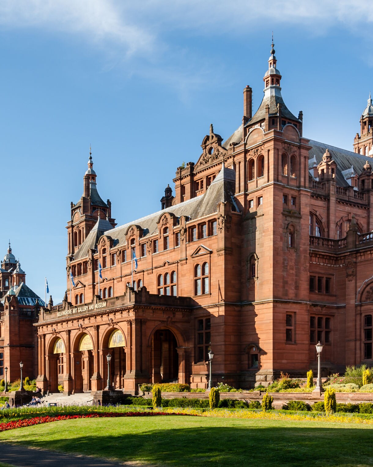 The red sandstone Kelvingrove Art Gallery and Museum in Glasgow with ornate towers, arched entrance and landscaped gardens under a clear blue sky.