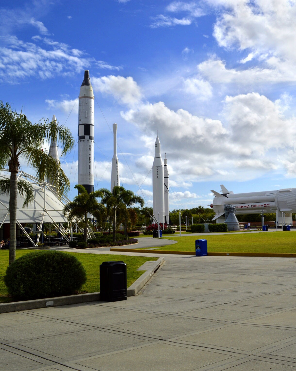 Outdoor rocket garden at Kennedy Space Center with several upright rockets and a massive Saturn V rocket displayed horizontally under a partly cloudy Florida sky.