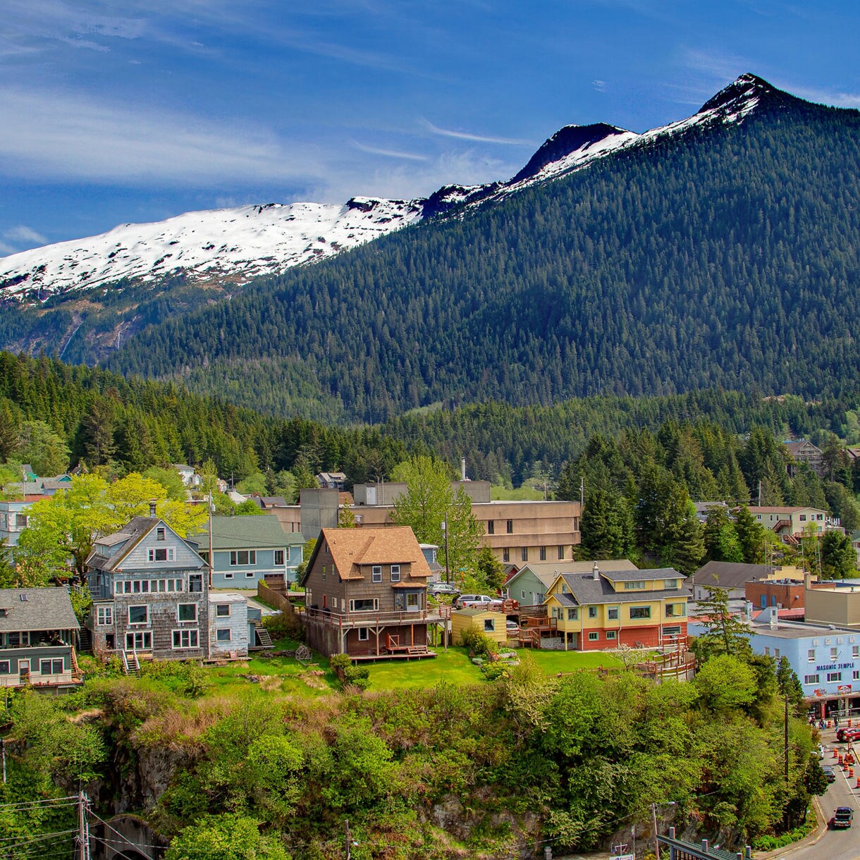 Aerial view of downtown Ketchikan, Alaska, with colorful historic buildings and homes nestled among lush green trees, backed by a massive snow-capped mountain under a bright blue sky.