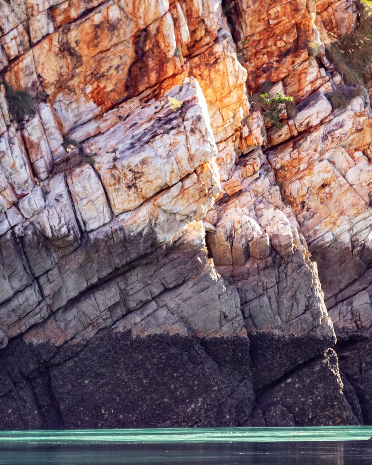Close-up view of layered Kimberley sandstone cliffs in warm red, orange and cream tones rising above still turquoise water.