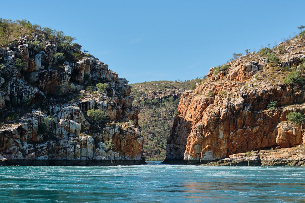 View of a narrow passage between tall, rugged sandstone cliffs with swirling turquoise water in the foreground and a sunlit hillside in the distance.