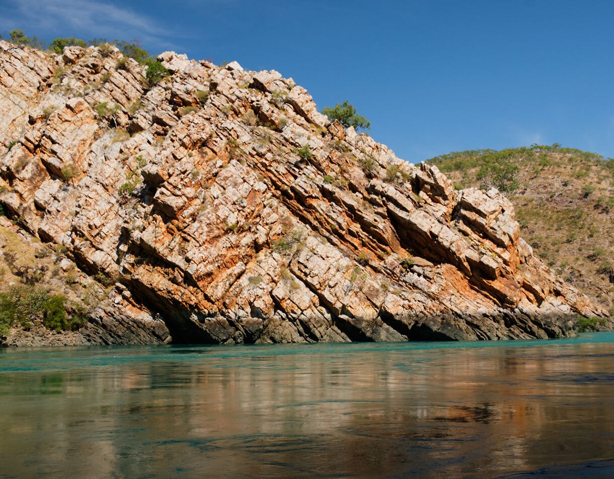 Sandstone cliff face with dramatic angled layers rising above calm turquoise water, with sunlit hills in the background under a clear blue sky.