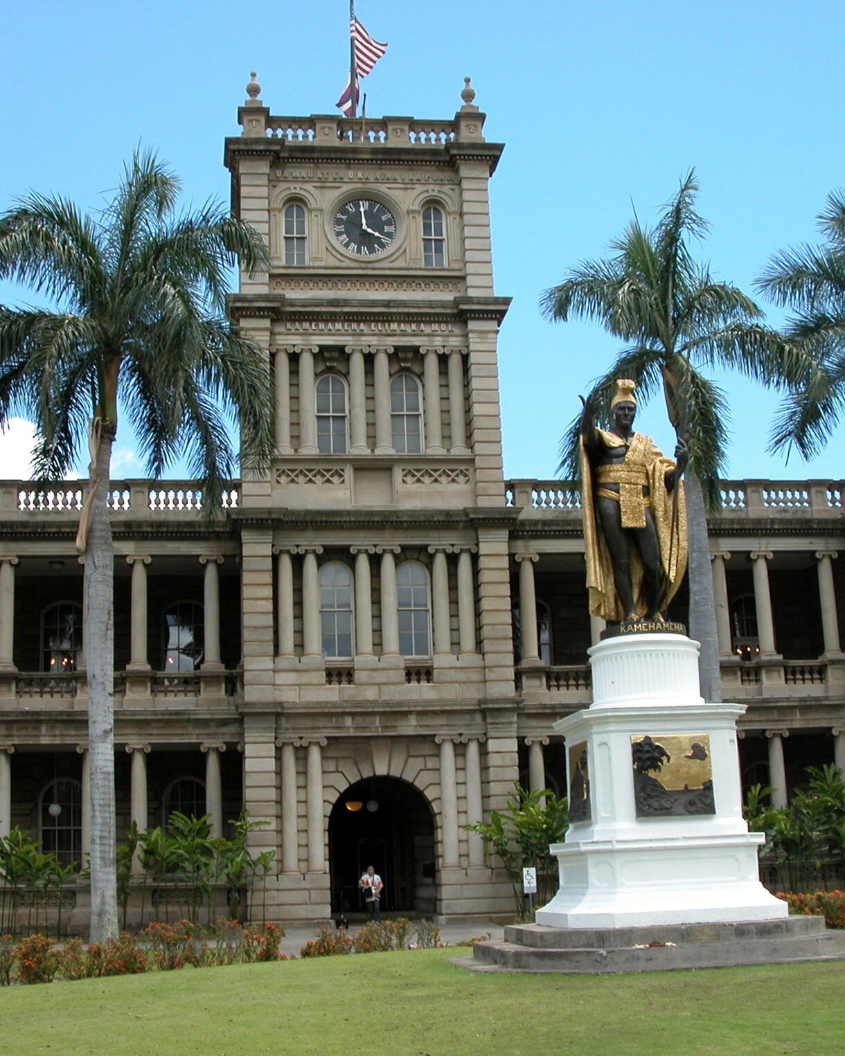 Bronze and gold statue of King Kamehameha I in front of Aliʻiōlani Hale, a historic courthouse with a clock tower and palm trees.