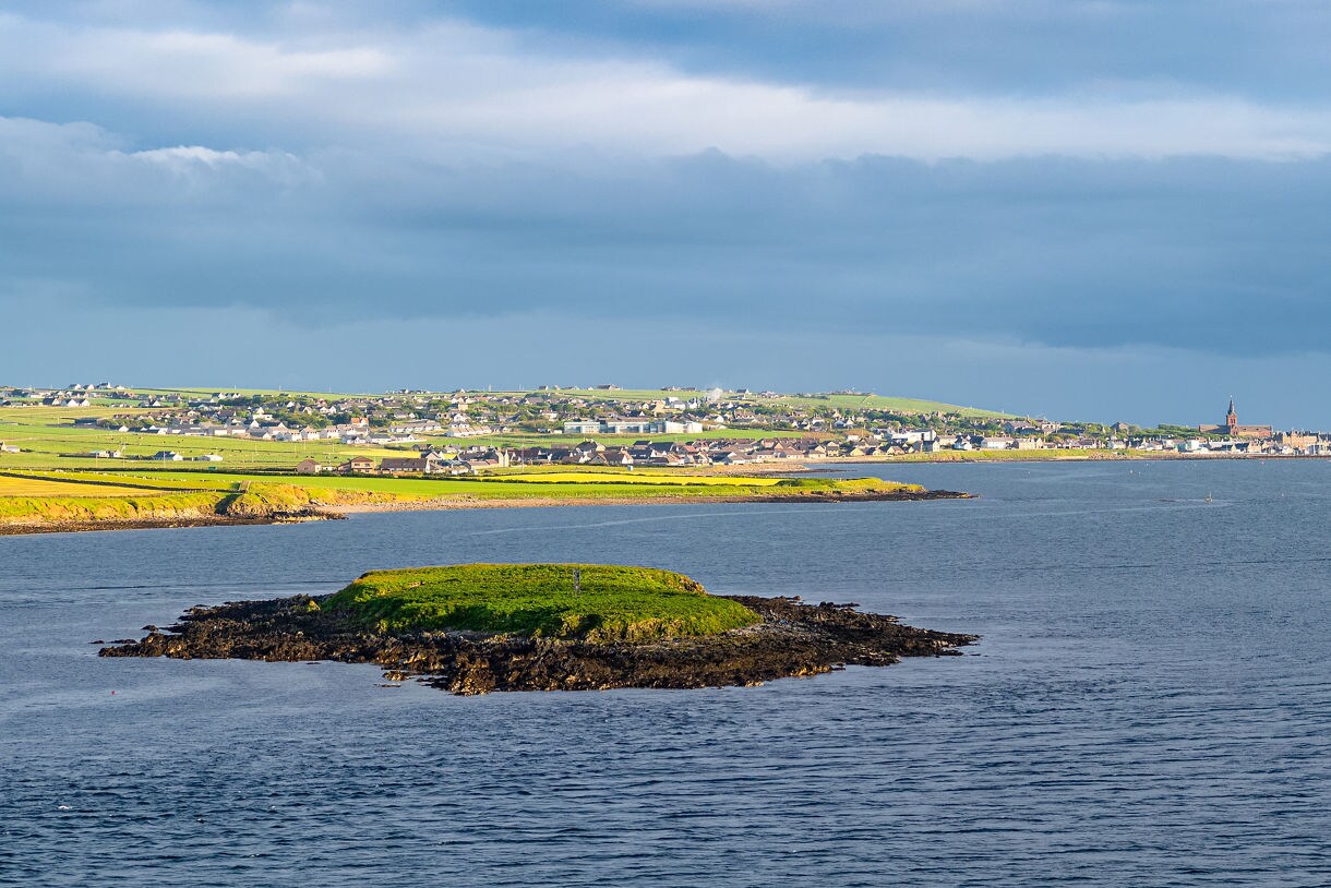 Coastal view of Kirkwall on the Orkney Isles, with green fields, scattered houses and the spire of St. Magnus Cathedral visible across the water.