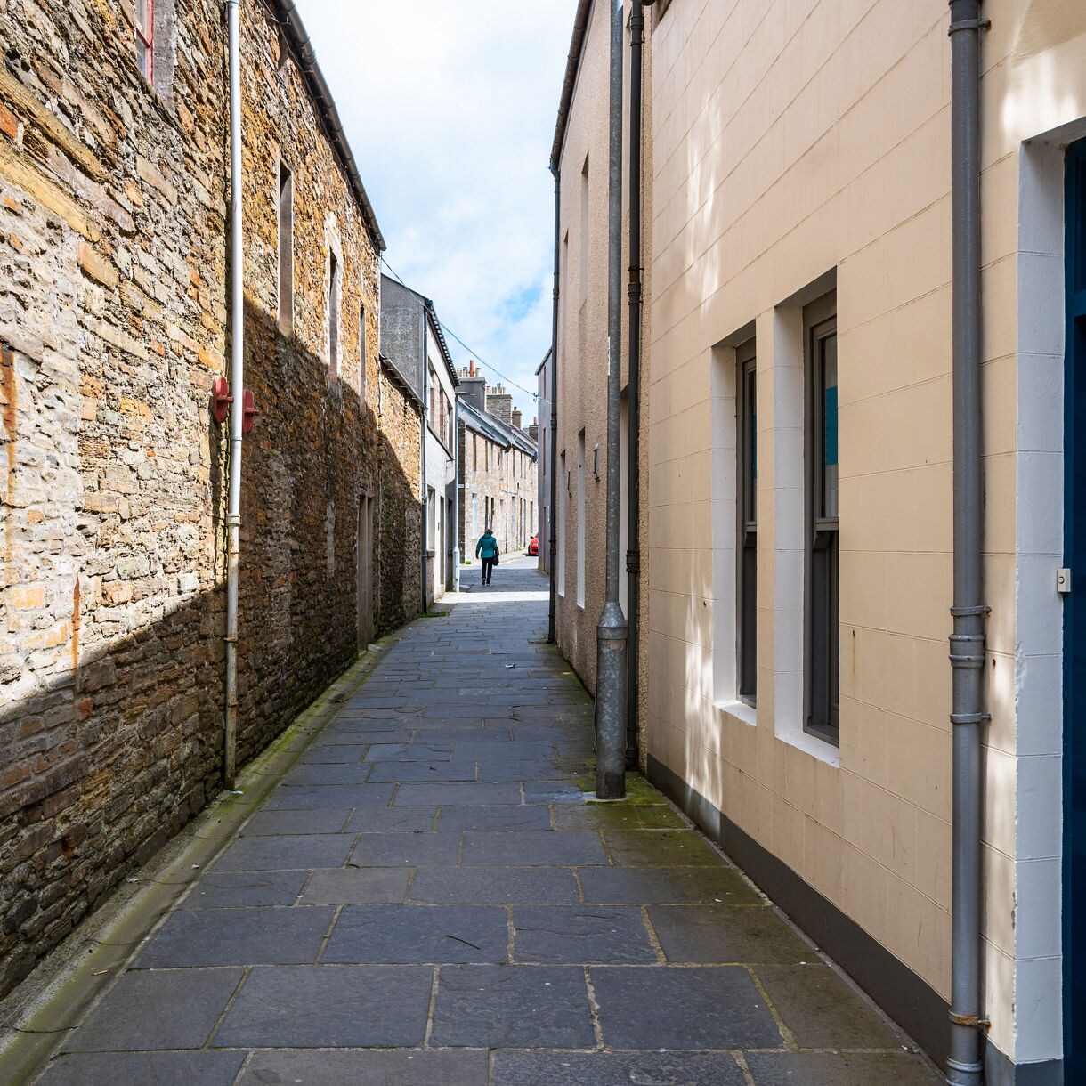 Narrow stone-paved alley in Kirkwall, Orkney, bordered by rustic stone walls and pastel buildings with a lone person walking in the distance.