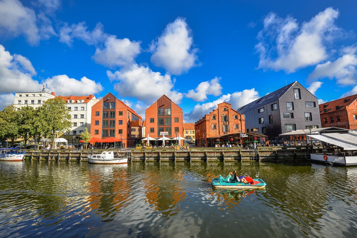 Red-brick and modern black gabled buildings along Klaipėda’s waterfront, with boats and a pedal boat on the river.