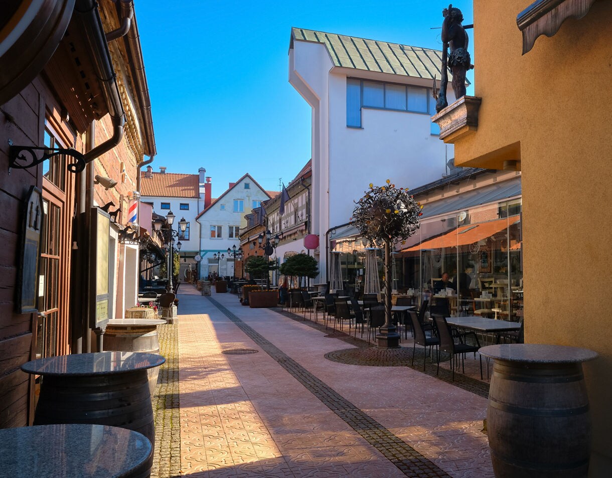 Narrow street in Klaipėda Old Town, lined with cafés, outdoor seating and historic buildings under a clear blue sky.