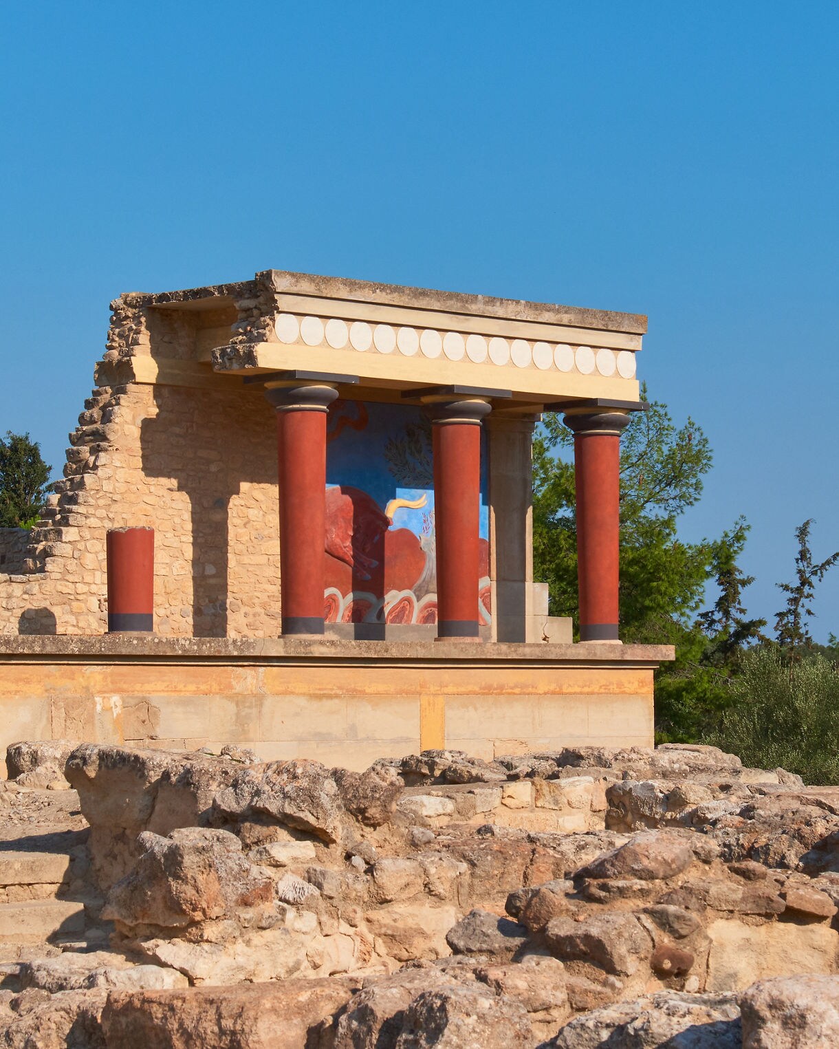 The restored ruins of the Palace of Knossos in Crete, featuring red columns, stone walls and colorful frescoes against a clear blue sky.
