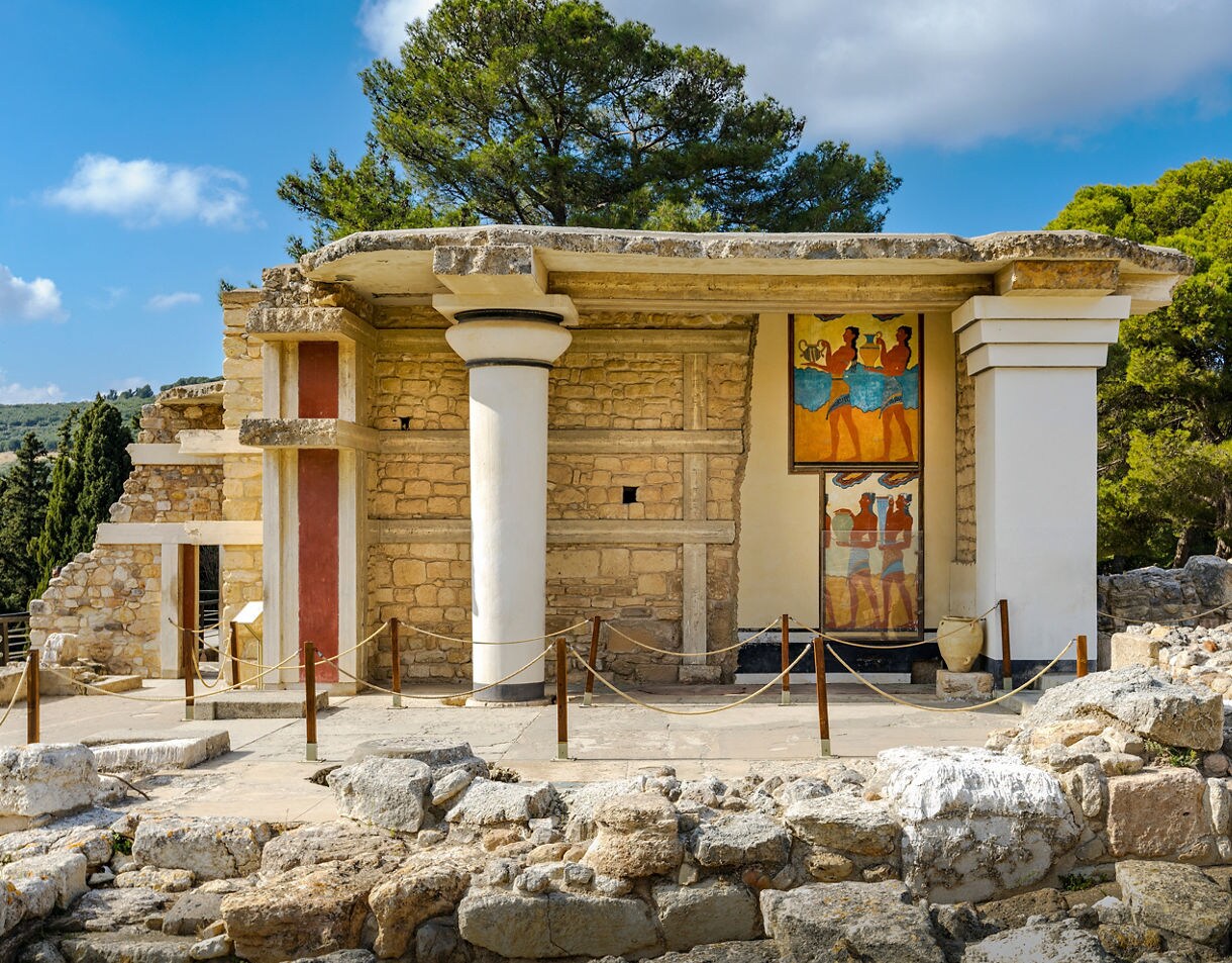 Restored section of the Palace of Knossos in Crete featuring stone walls, white columns with red accents and a vivid fresco of Minoan figures, surrounded by rocky ruins and lush green trees under a bright sky.