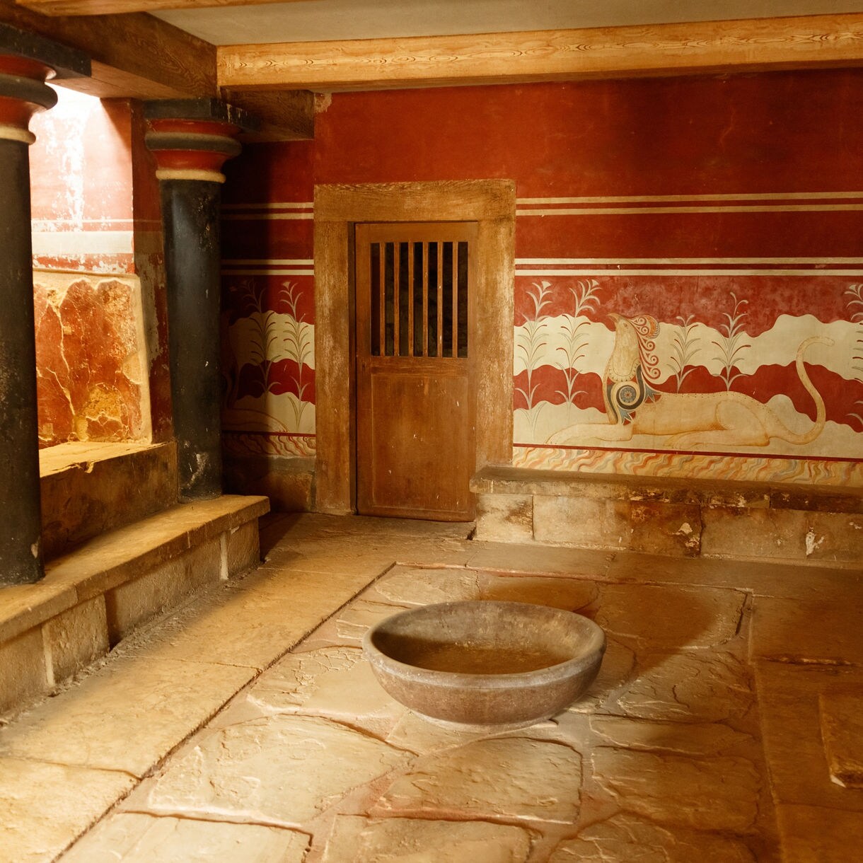 Interior chamber of the Palace of Knossos showing restored red walls, painted frescoes with griffin motifs, black-and-red columns and a stone basin set on the floor.