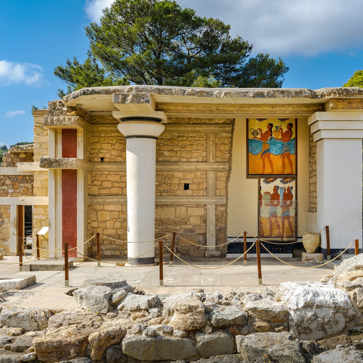 Restored section of the Palace of Knossos featuring red and white Minoan columns, tan stone walls and colorful frescoes of human figures, surrounded by scattered ancient ruins and framed by trees and a bright blue sky.