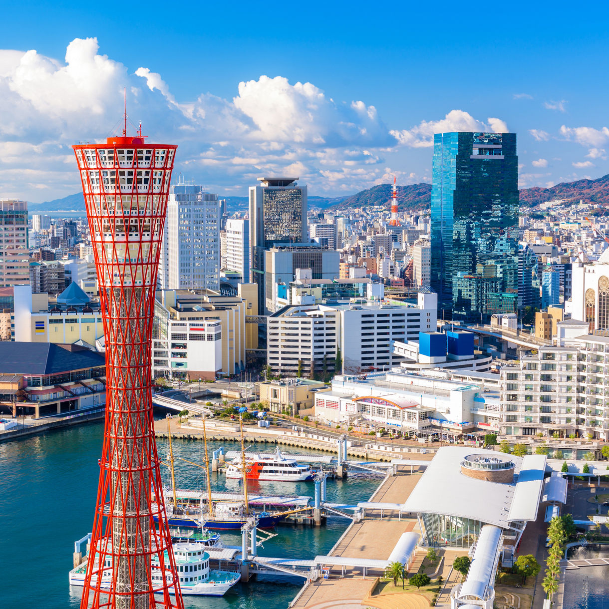 Red lattice-frame Kobe Port Tower rising above the harbor with docked boats, waterfront buildings and city skyline behind it and mountains in the distance.