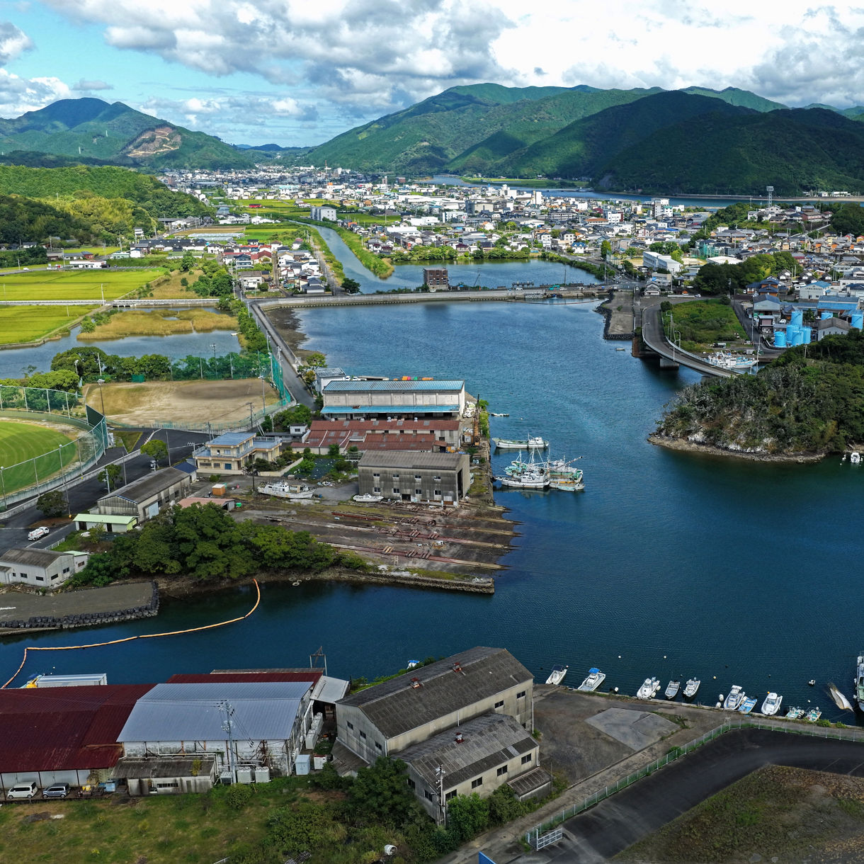 An elevated aerial view looking down at a calm harbor inlet filled with moored fishing boats, bounded by coastal buildings, green fields and rolling mountains under a bright sky with fluffy clouds.