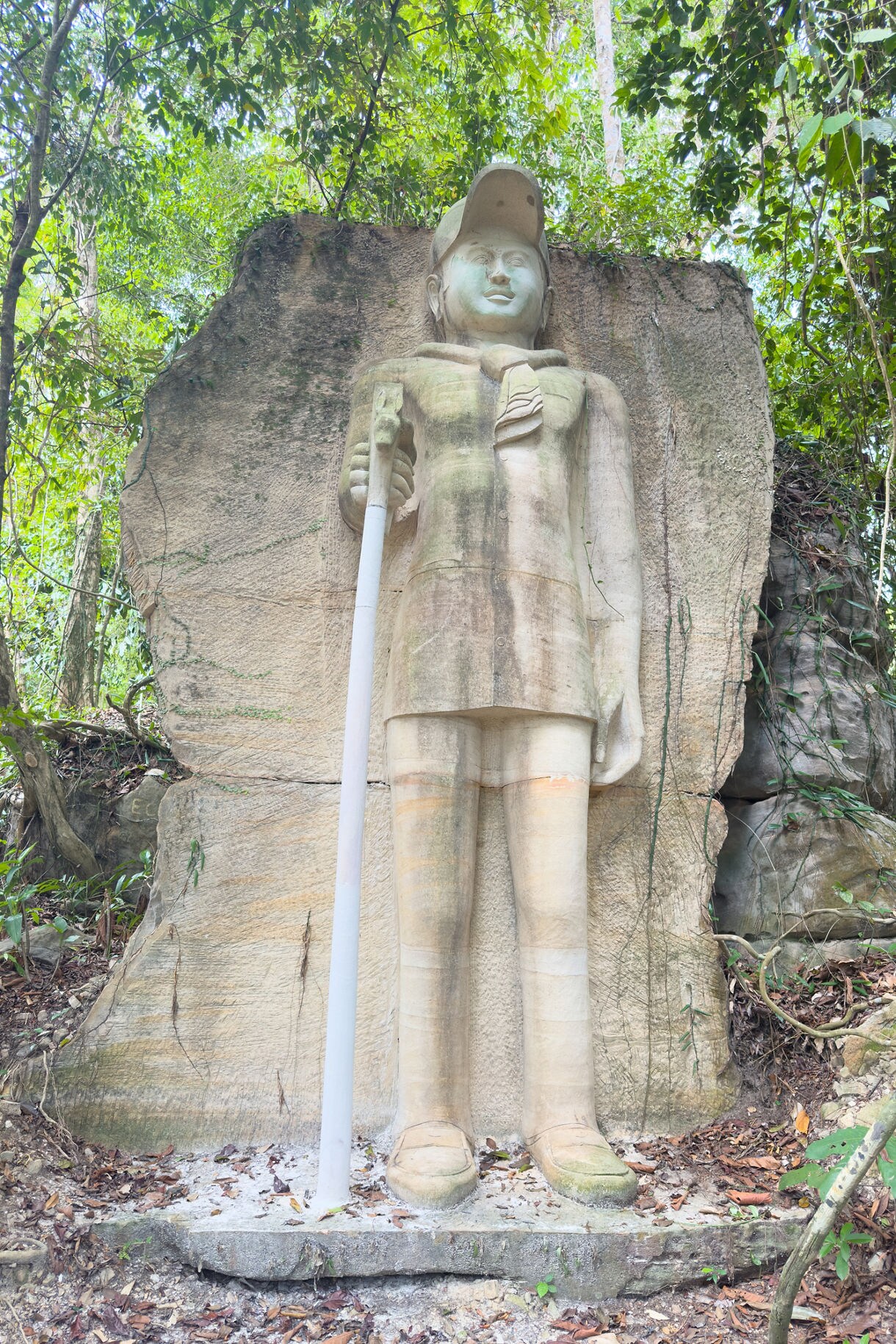 Tall carved stone statue of a person in boots holding a staff, set against a rock slab and surrounded by dense green forest.