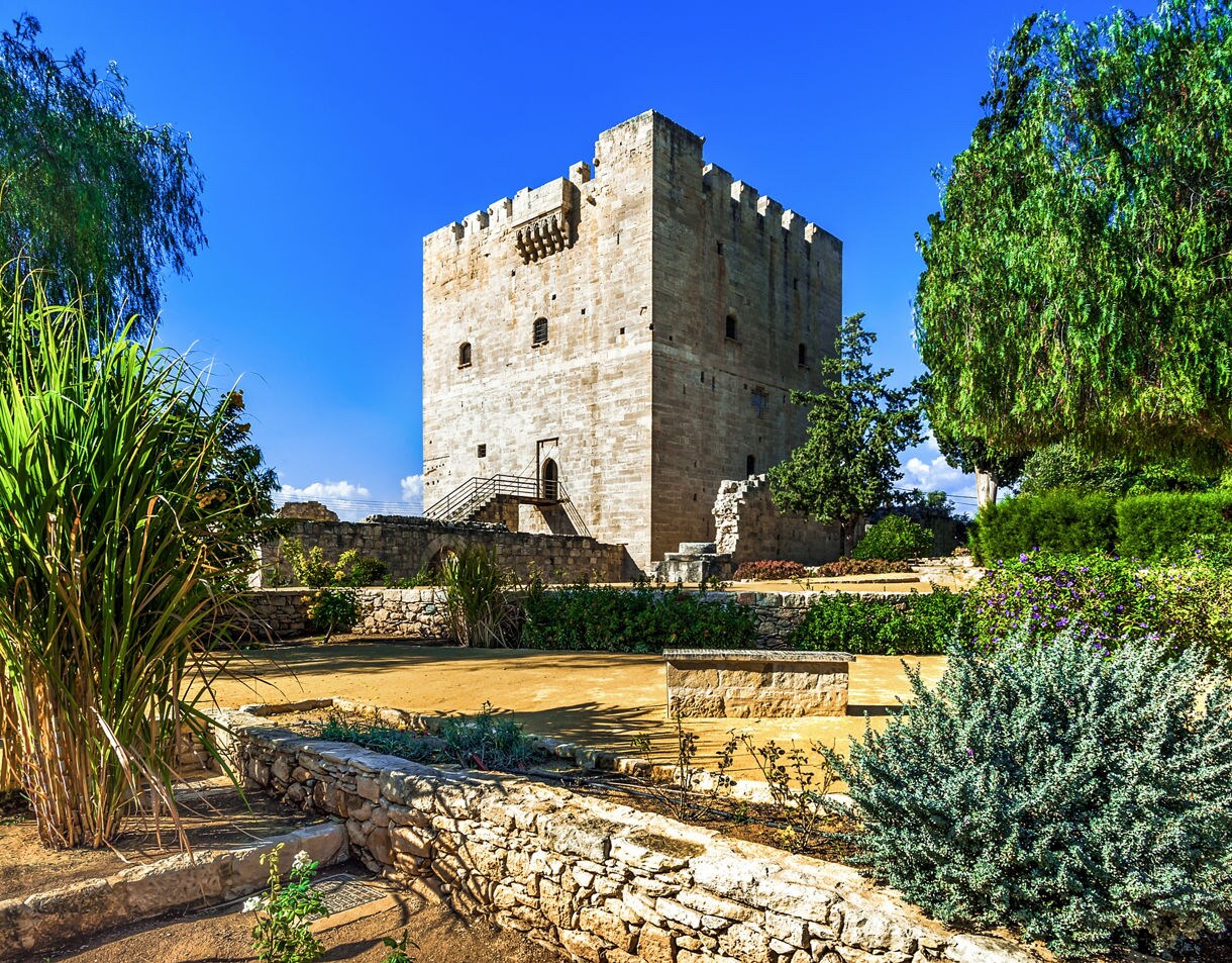 Stone Crusader-era Kolossi Castle rises behind lush Mediterranean greenery under a bright blue sky, featuring a square tower, battlements and surrounding garden paths.