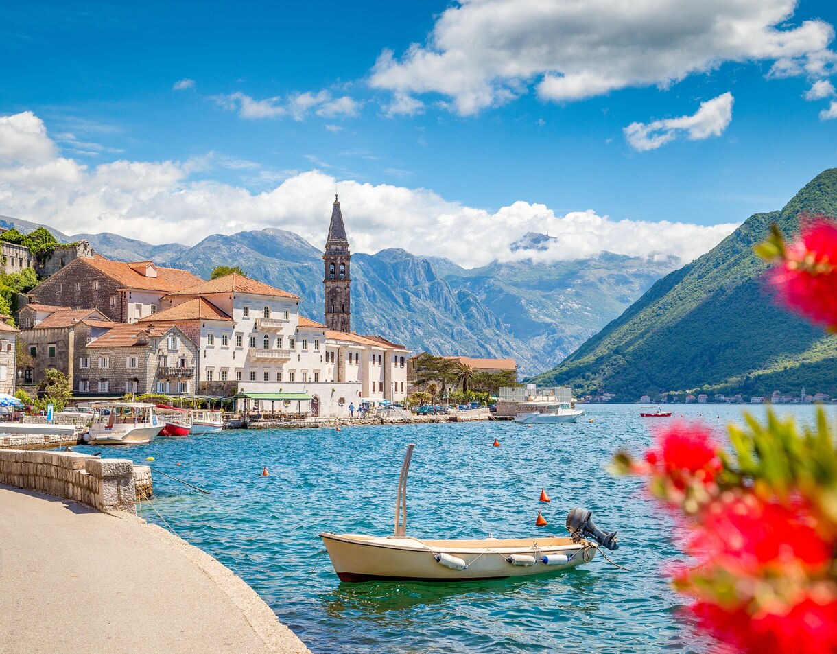 A small boat floating on turquoise water along Perast’s historic waterfront, with stone buildings and mountains in the background.